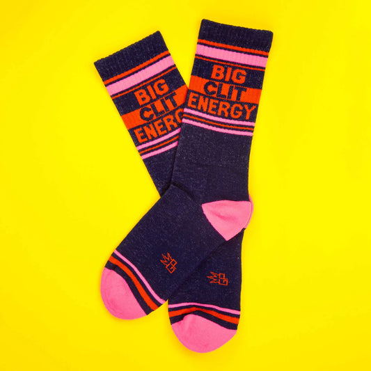 Navy and pink socks featuring 'Big Clit Energy' in bright lettering, arranged side-by-side on a vibrant yellow backdrop.