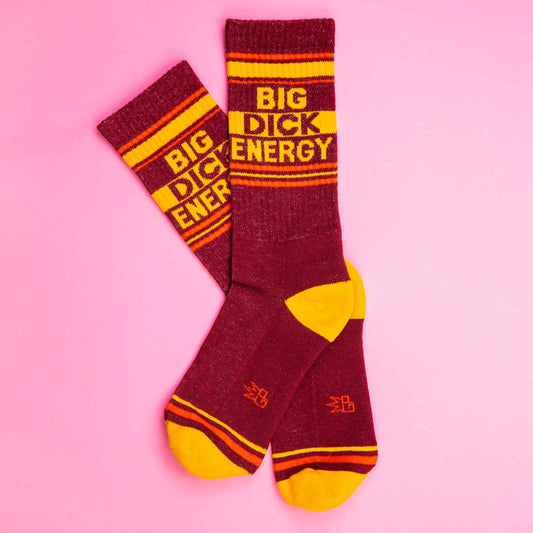 Crossed maroon and yellow socks laid on a pink background, featuring the phrase 'Big Dick Energy' in retro-style bold text.