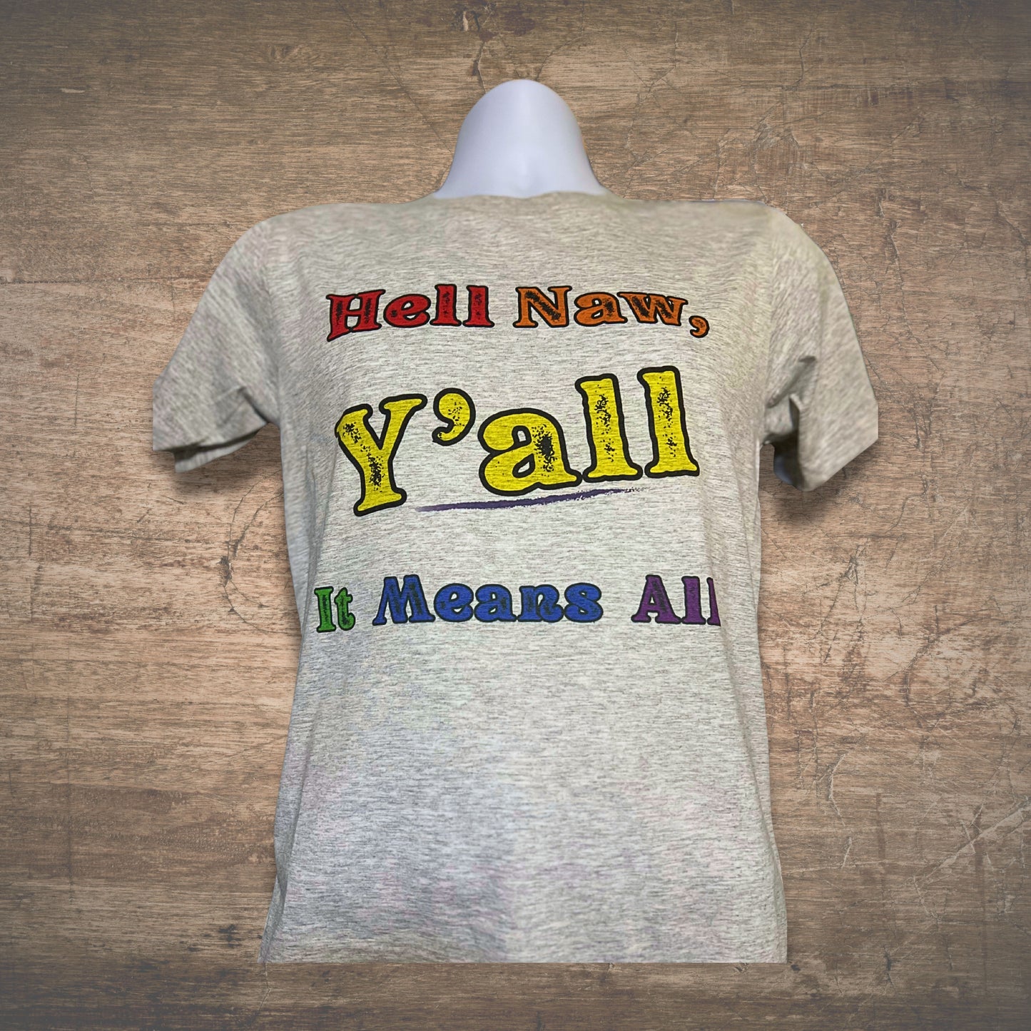 Gray t-shirt with colorful text, Hell Naw Y'all It Means All, on a wooden surface