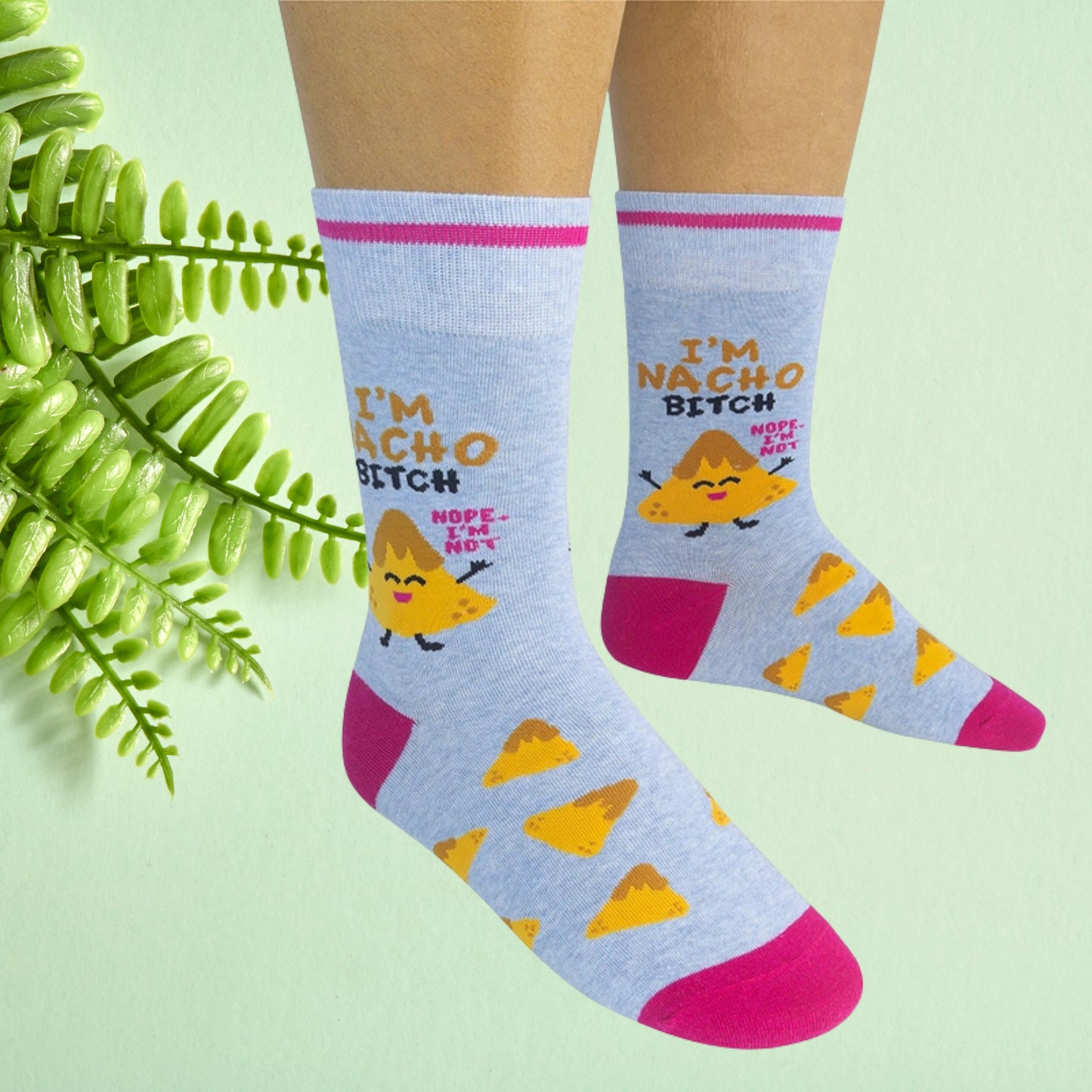Light blue crew socks with nacho chip character and “I’m nacho bitch” text, worn on feet against a soft green background.