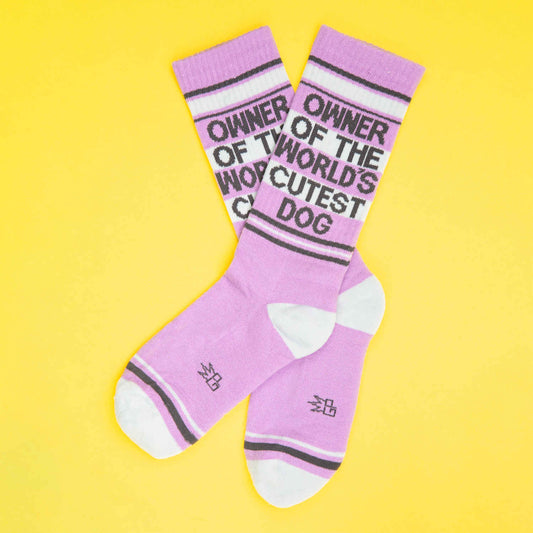 Playful pink and white novelty socks with black lettering saying "Owner of the World's Cutest Dog," photographed on a bright yellow background.