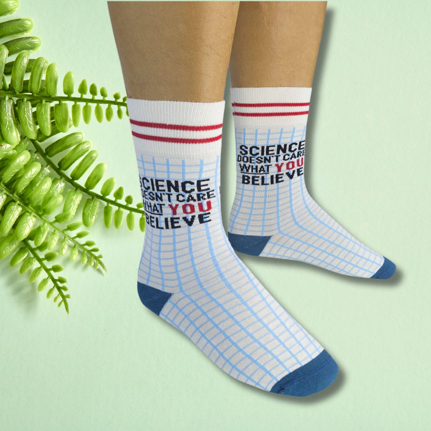 White crew socks with blue grid pattern and “Science doesn’t care what you believe” text, worn on feet against a soft green background.