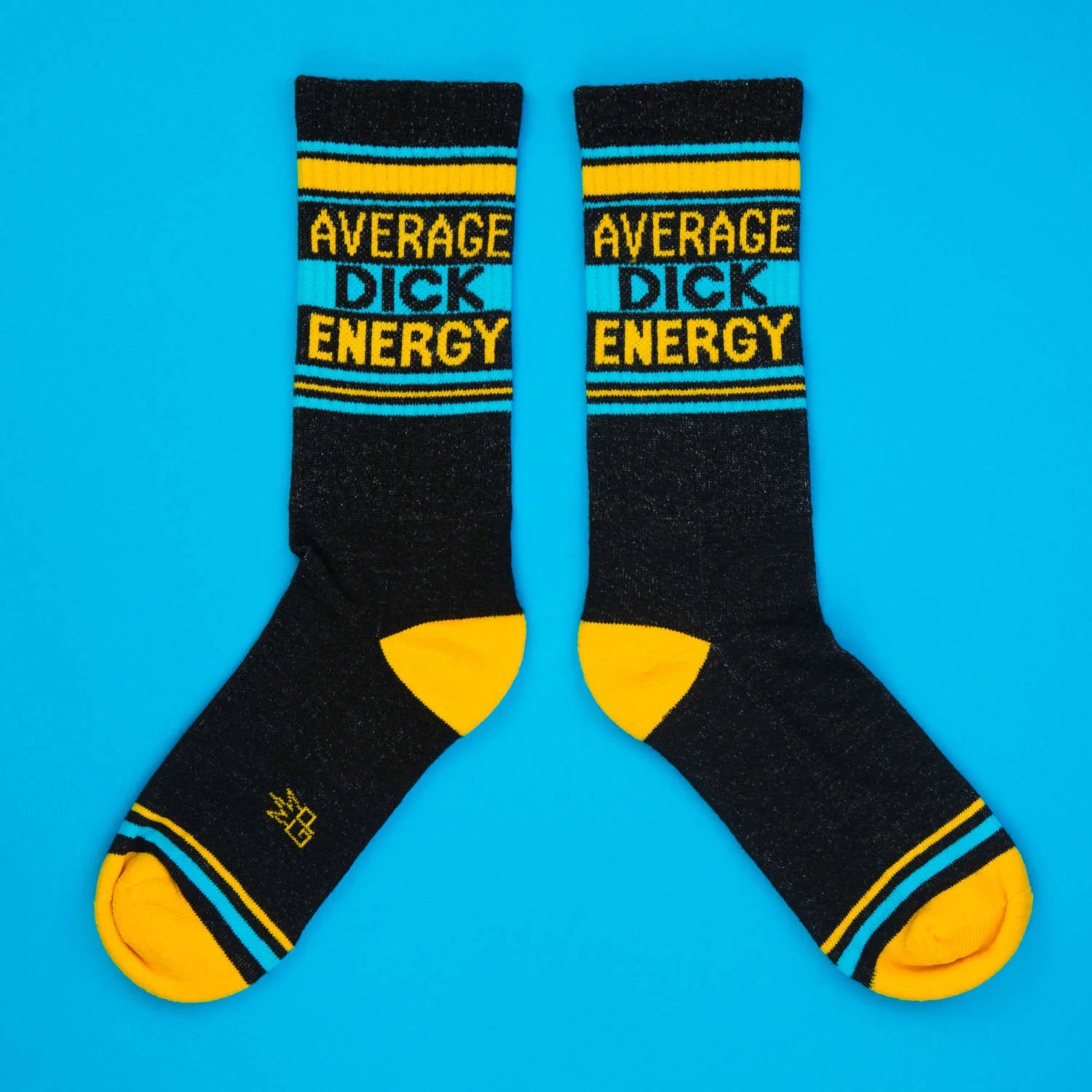 Black crew socks with yellow toe and heel, turquoise stripes, and bold text that reads Average Dick Energy. Flat lay on blue.