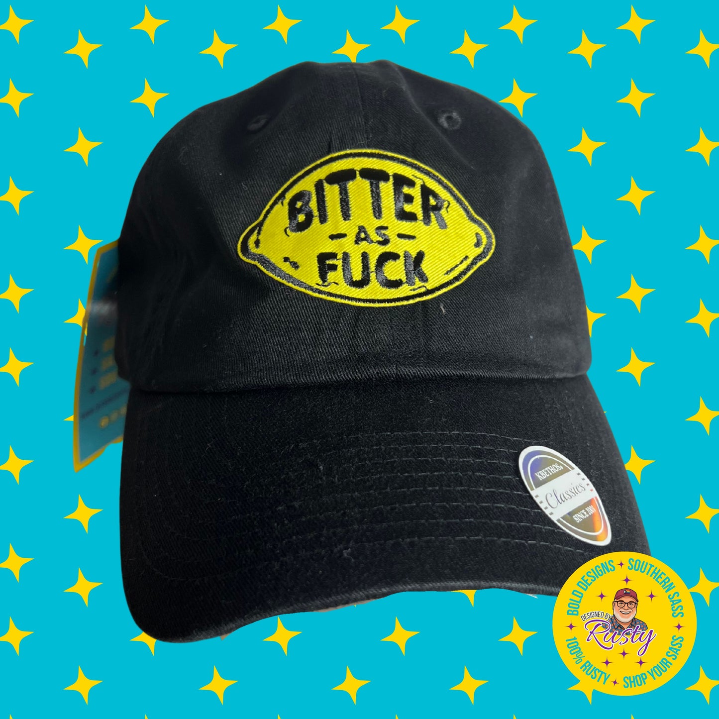 Black embroidered patch hat with yellow lemon design and bold text reading “Bitter As Fuck,” on turquoise background with yellow stars and Designed by Rusty logo.