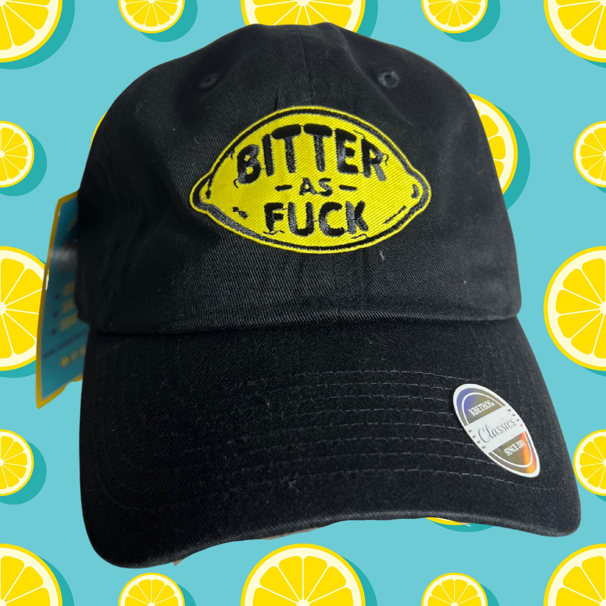 Black embroidered patch hat with yellow lemon design and bold text reading “Bitter As Fuck,” displayed against turquoise background with repeating lemon pattern.