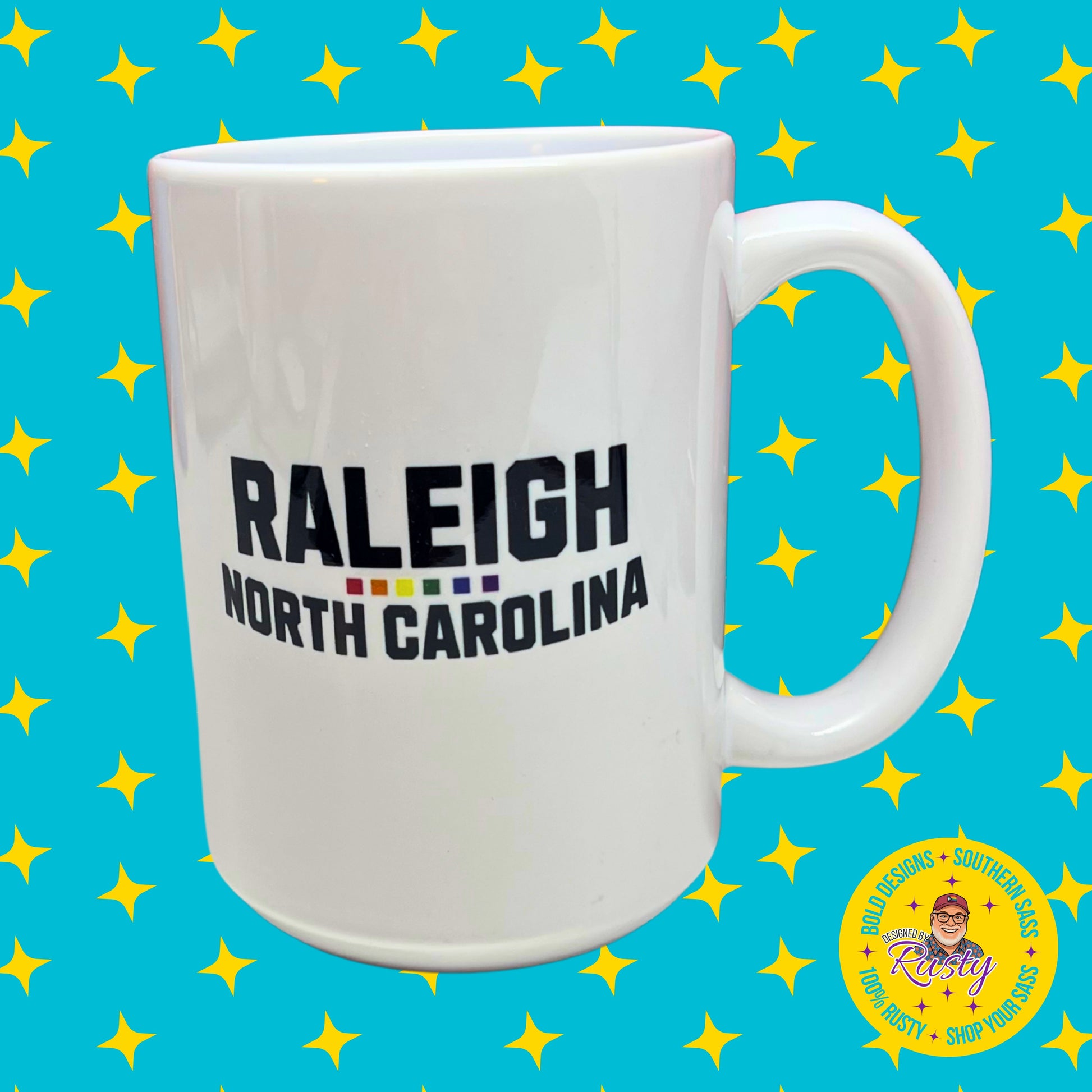 White ceramic 15 oz mug with bold black text “Raleigh North Carolina” and rainbow underline, on turquoise background with yellow stars and Designed by Rusty logo.