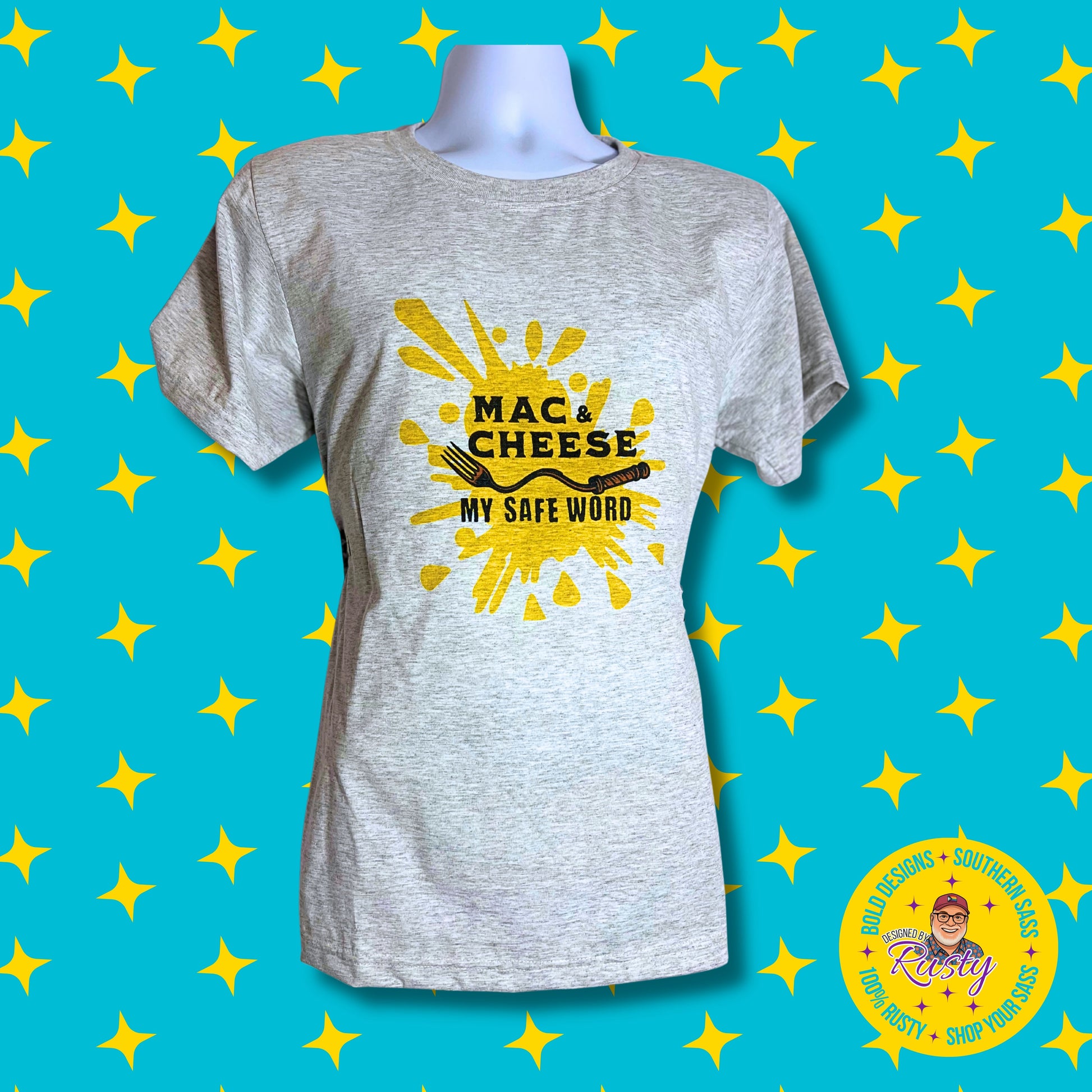 Light gray T-shirt with bold yellow splash design and text “Mac & Cheese My Safe Word,” on turquoise background with yellow stars and Designed by Rusty logo.