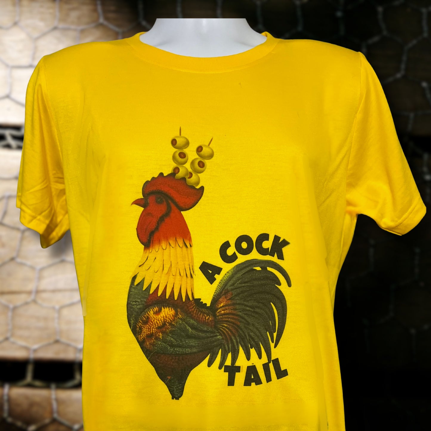 Yellow T-shirt with black rooster illustration wearing olives on toothpicks as a headpiece and the word Cocktail, displayed against rustic chicken wire background.
