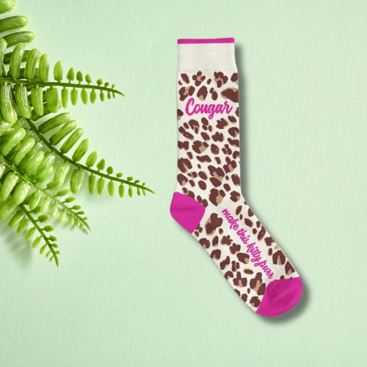 Leopard print crew socks with pink accents and “Cougar” text worn on feet against a soft green background.