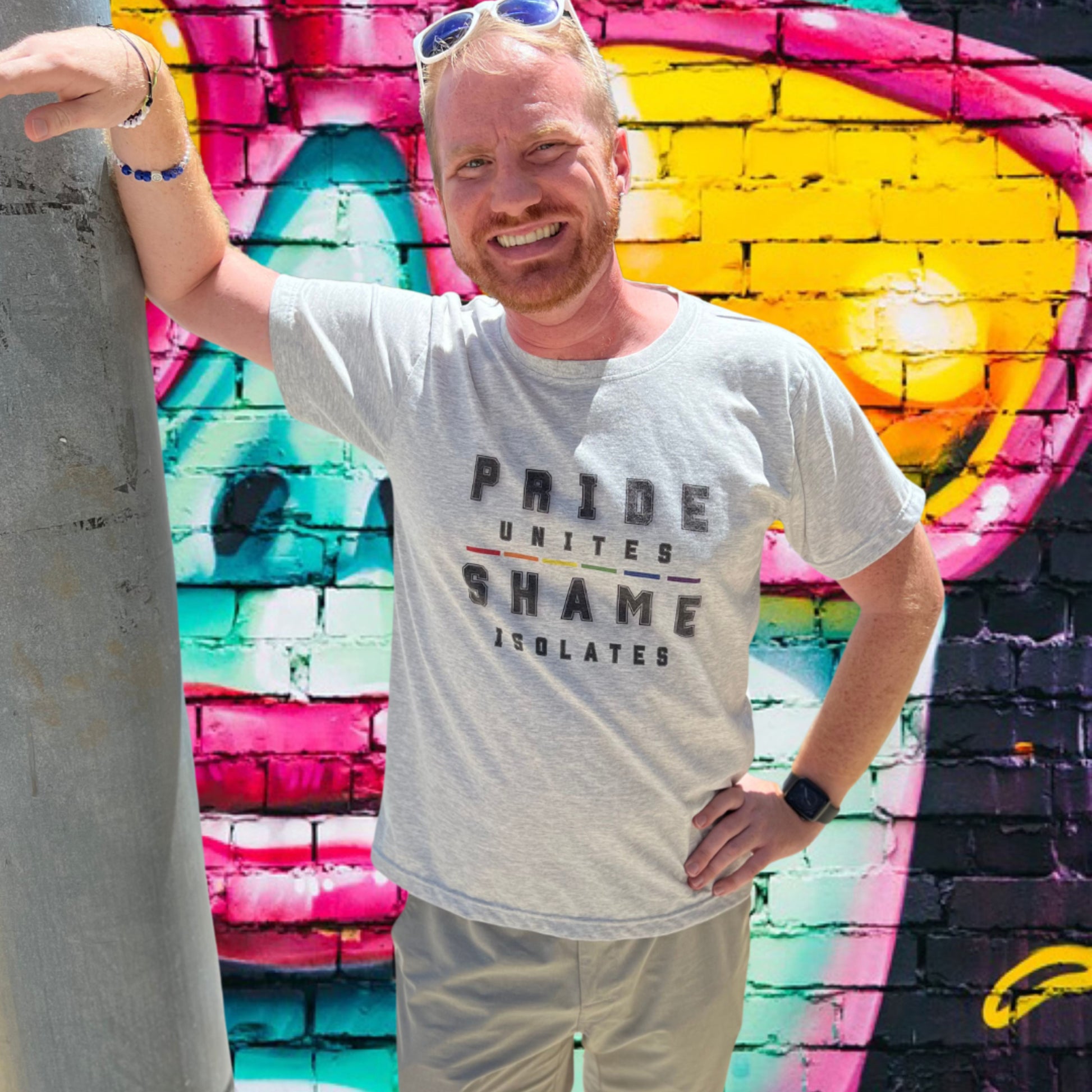 Man wearing a 'Pride Unites Shame Isolates' t-shirt in front of a colorful mural.