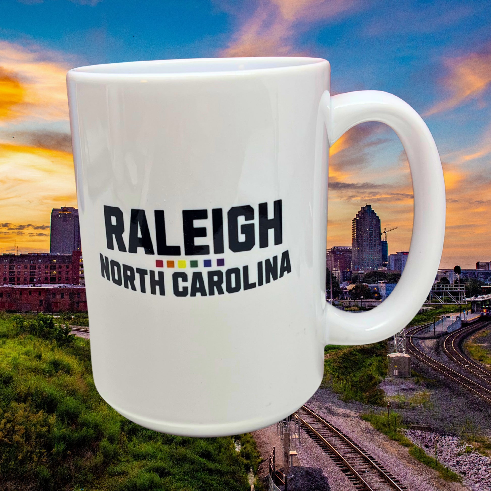 White ceramic 15 oz mug with bold black text “Raleigh North Carolina” and rainbow underline, displayed against Raleigh city skyline at sunset.