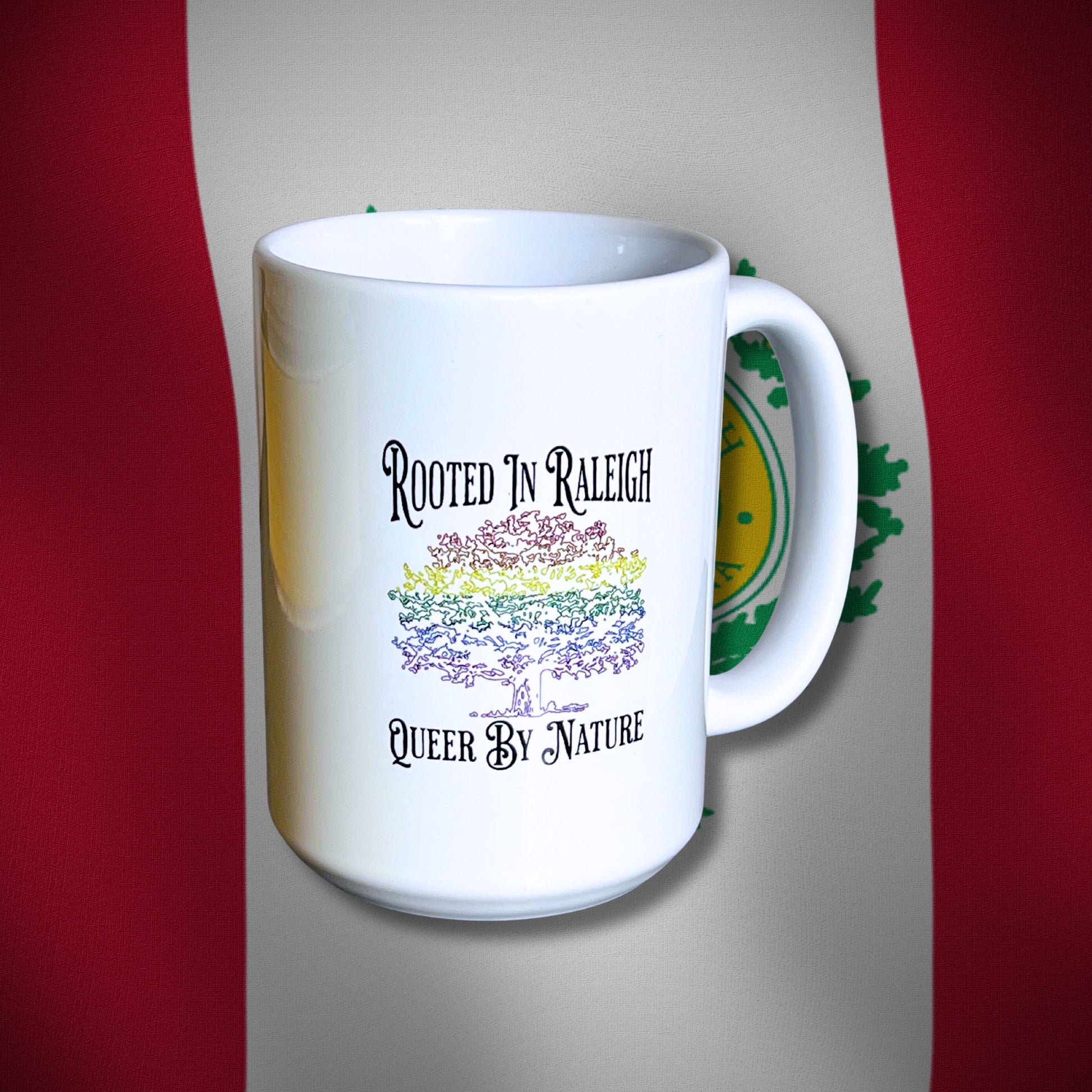 White 15 oz ceramic mug with rainbow oak tree design and Rooted in Raleigh text, displayed in front of Raleigh city flag background.