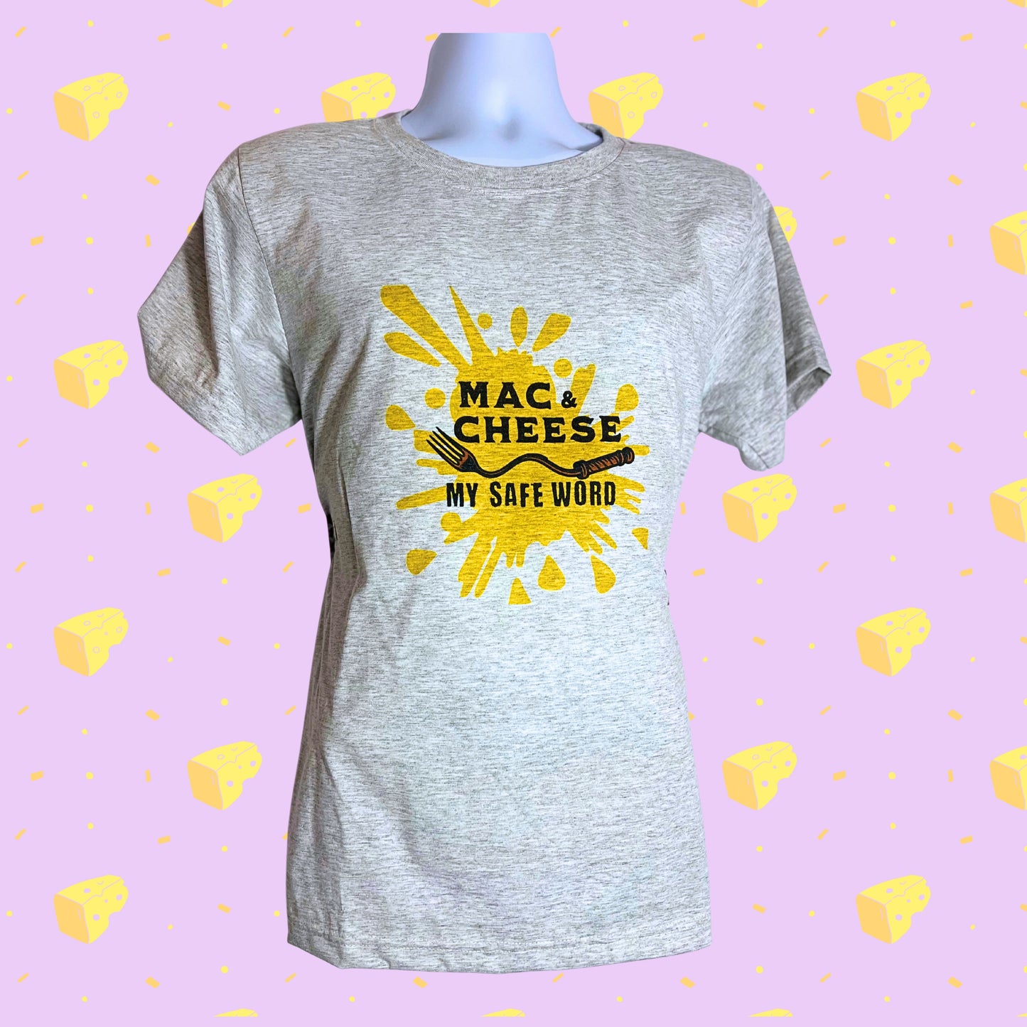 Light gray T-shirt with bold yellow splash design and text “Mac & Cheese My Safe Word,” displayed against a pastel purple background with scattered cheese icons.