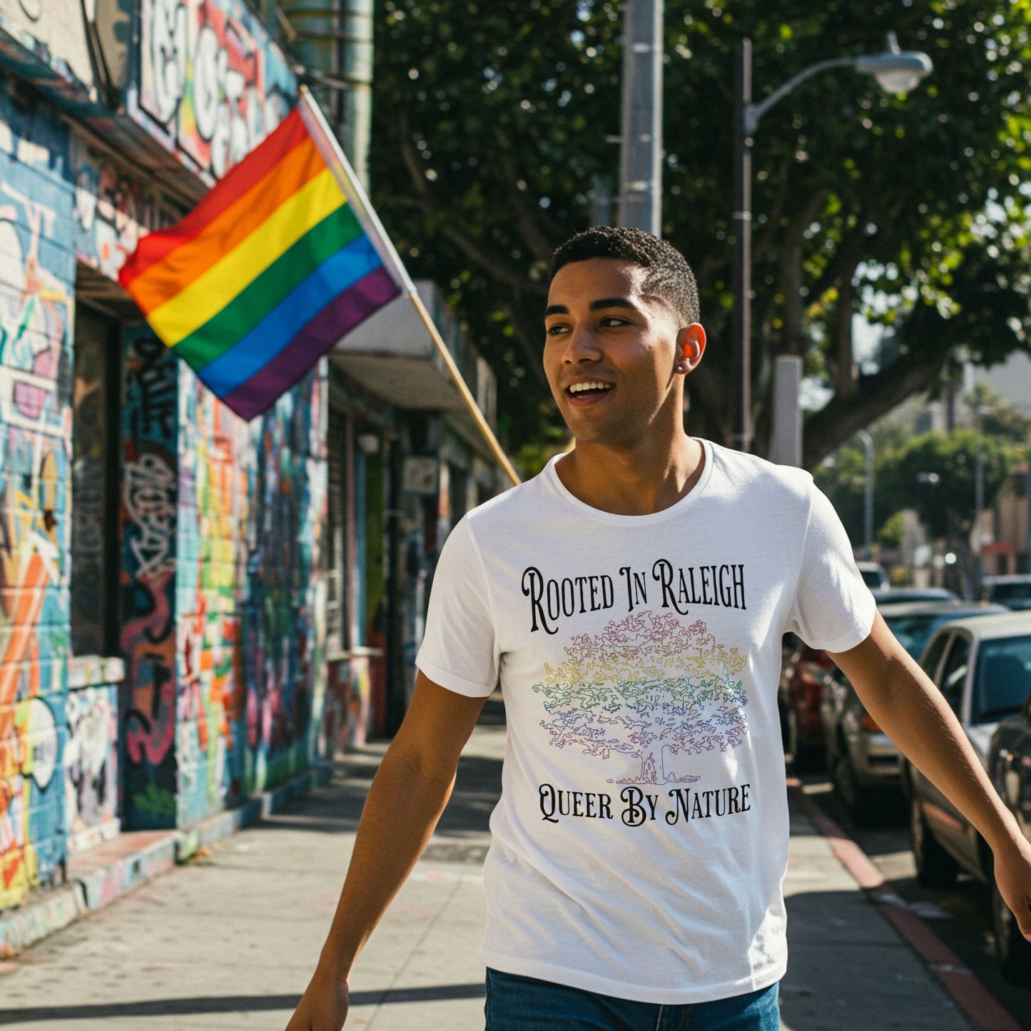 Person wearing light gray T-shirt with rainbow tree and “Rooted in Raleigh – Queer by Nature” text, walking on a city street with pride flag in background.