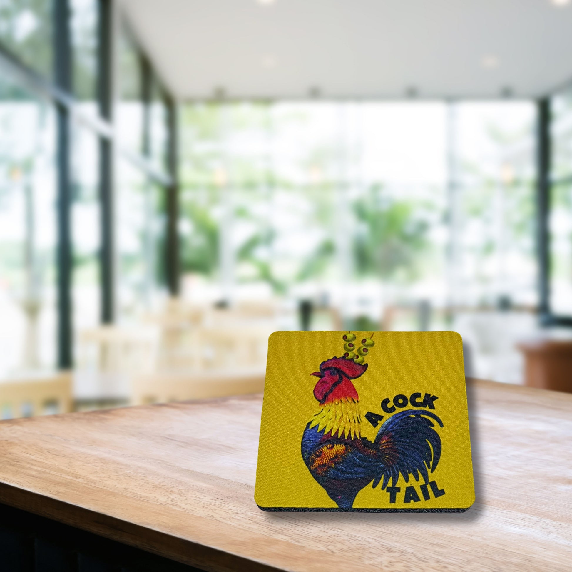 Yellow “A Cocktail” rooster coaster with martini olive garnish, displayed on a wooden café table with blurred background.
