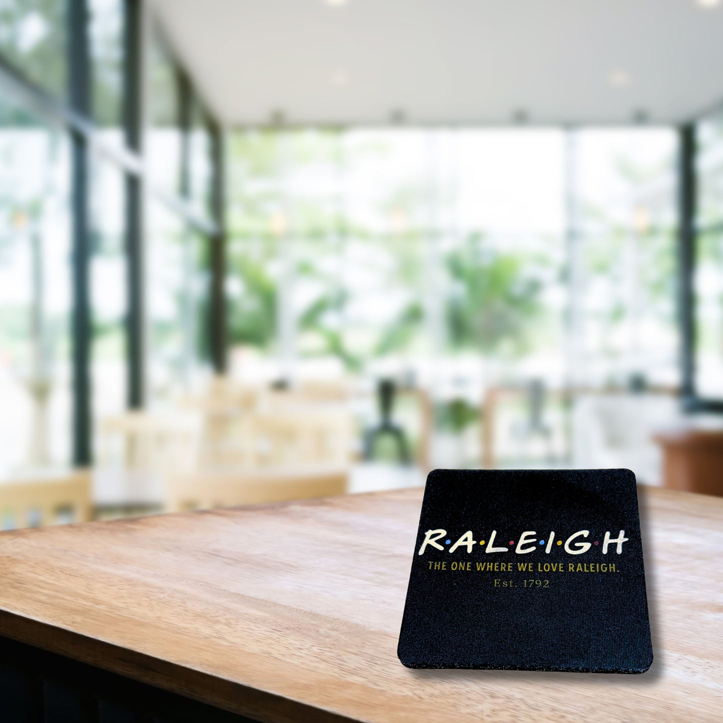 Raleigh Friends-inspired coaster on a wooden table with blurred café background, featuring text 'The one where we love Raleigh. Est. 1792.'