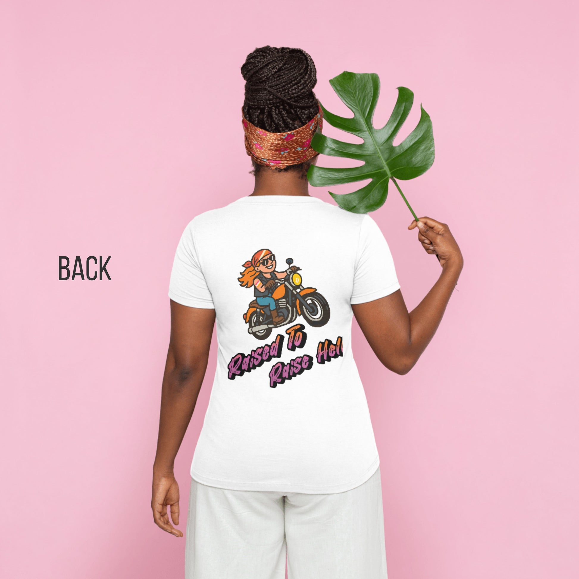 Woman modeling white T-shirt with cartoon biker woman graphic and “Raised to Raise Hell” text on the back, shown against a pink background holding a leaf.