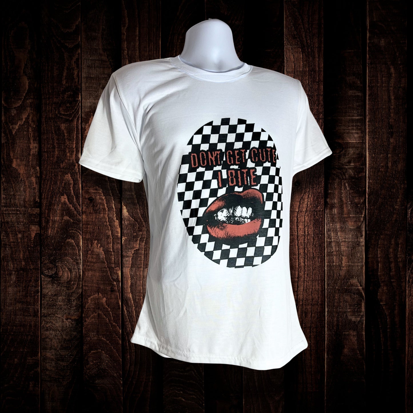 White T-shirt with bold design of red lips biting and text “Don’t Get Cute I Bite” over a black-and-white checkerboard oval, displayed on a mannequin against a dark wood background.