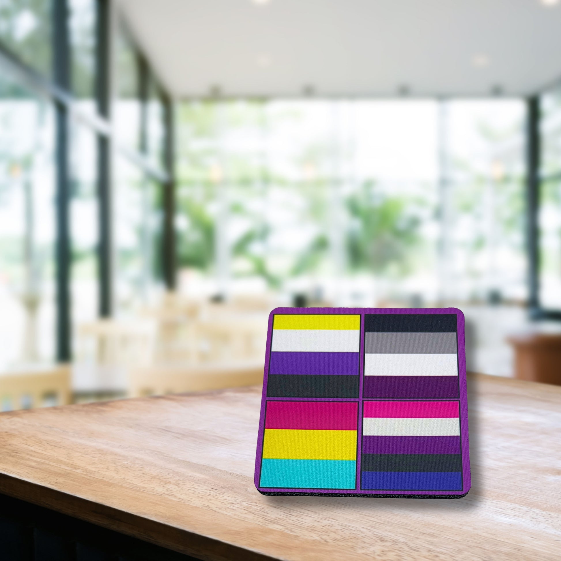 Pride flag coaster with purple border displayed on wooden café table in a bright modern setting.