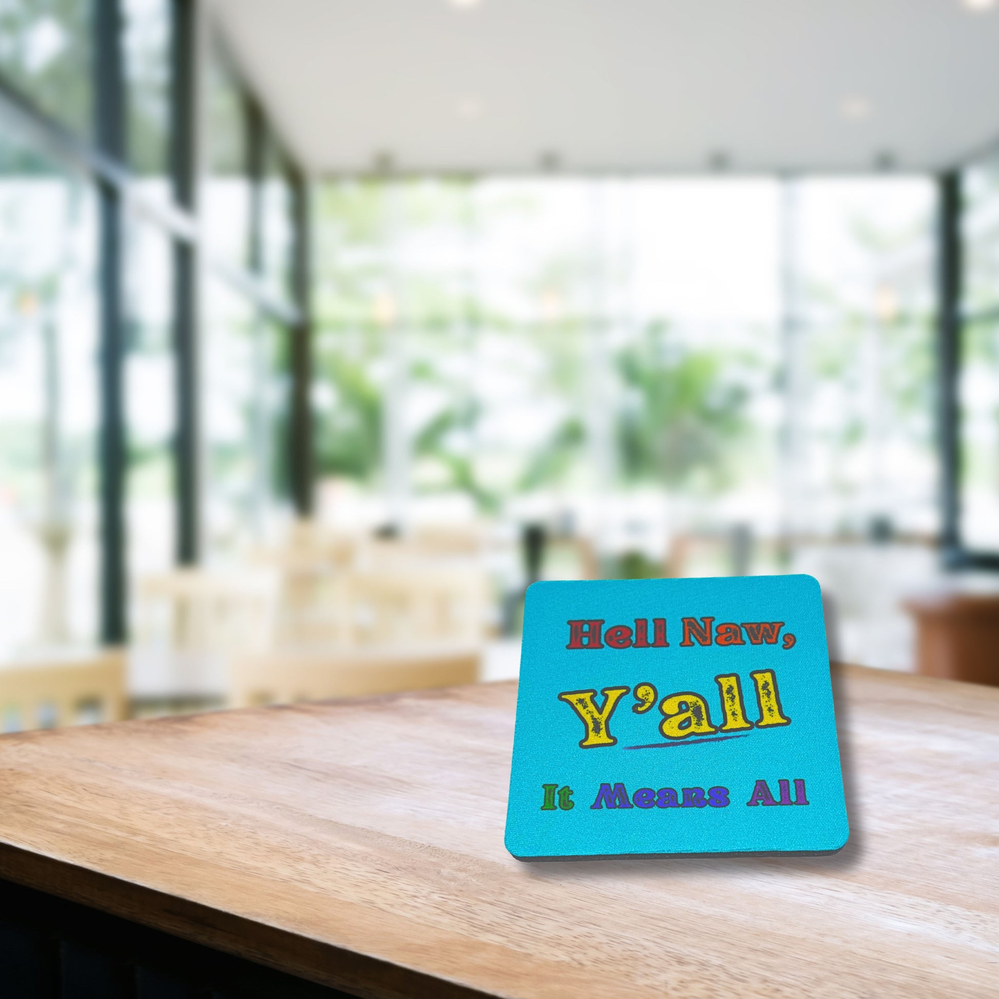 Funny Southern sass coaster reading “Hell Naw, Y’all It Means All,” placed on a wooden café table with blurred modern background.