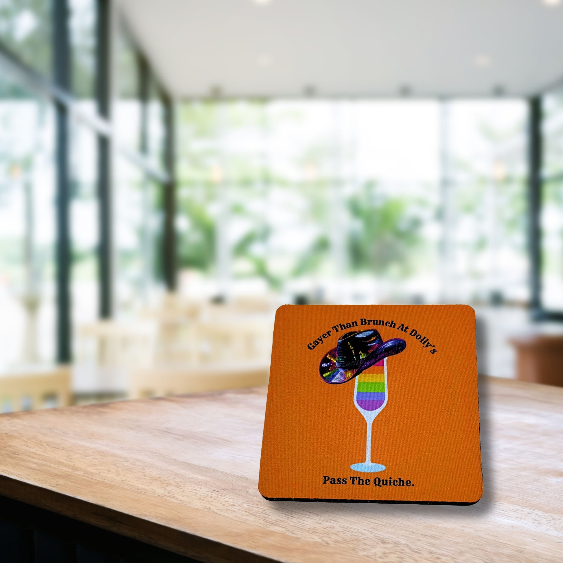 Orange coaster with rainbow mimosa glass and sequin cowboy hat reading “Gayer Than Brunch at Dolly’s – Pass The Quiche,” displayed on a wooden café table with bright blurred background.