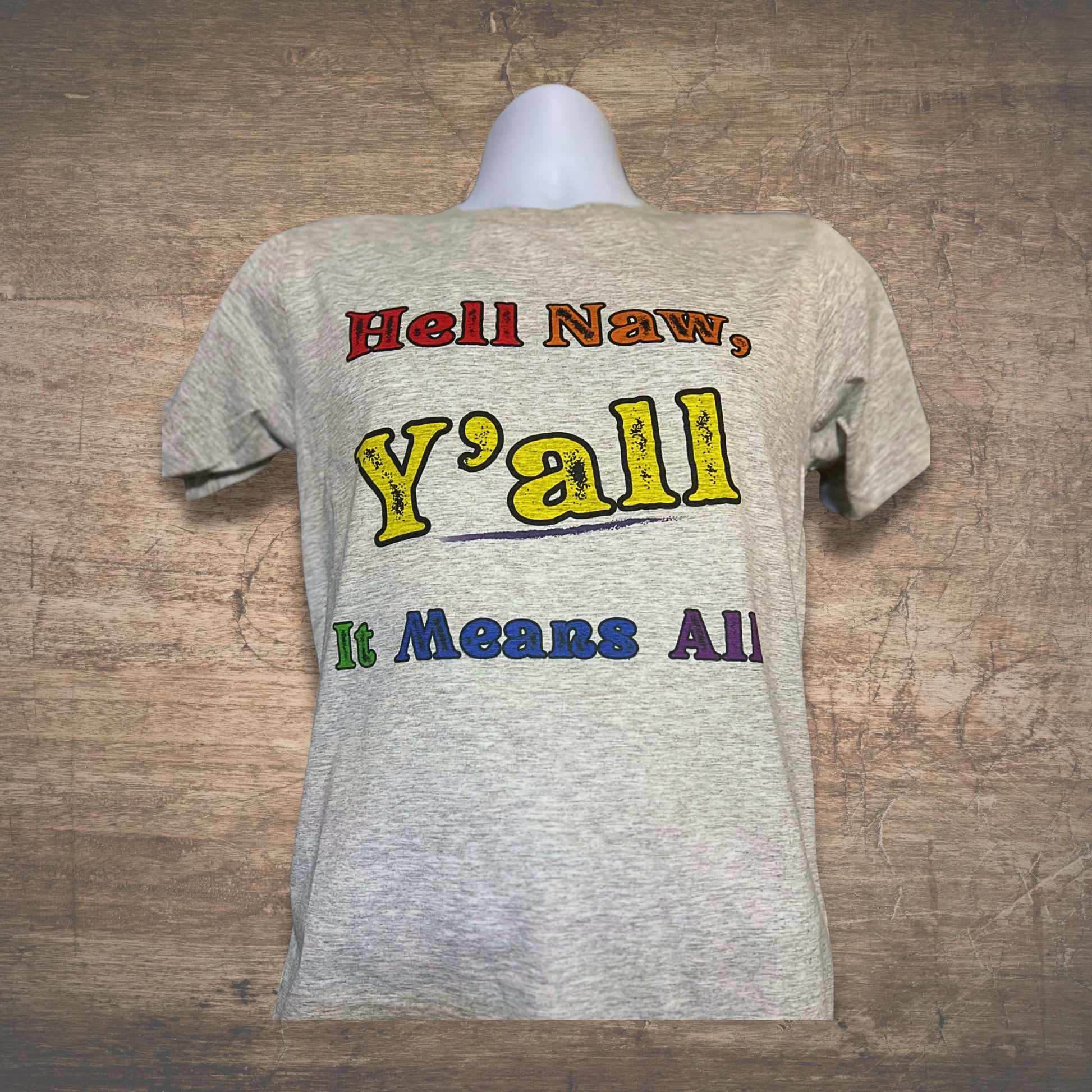 Gray t-shirt with colorful text, Hell Naw Y'all It Means All, on a wooden surface