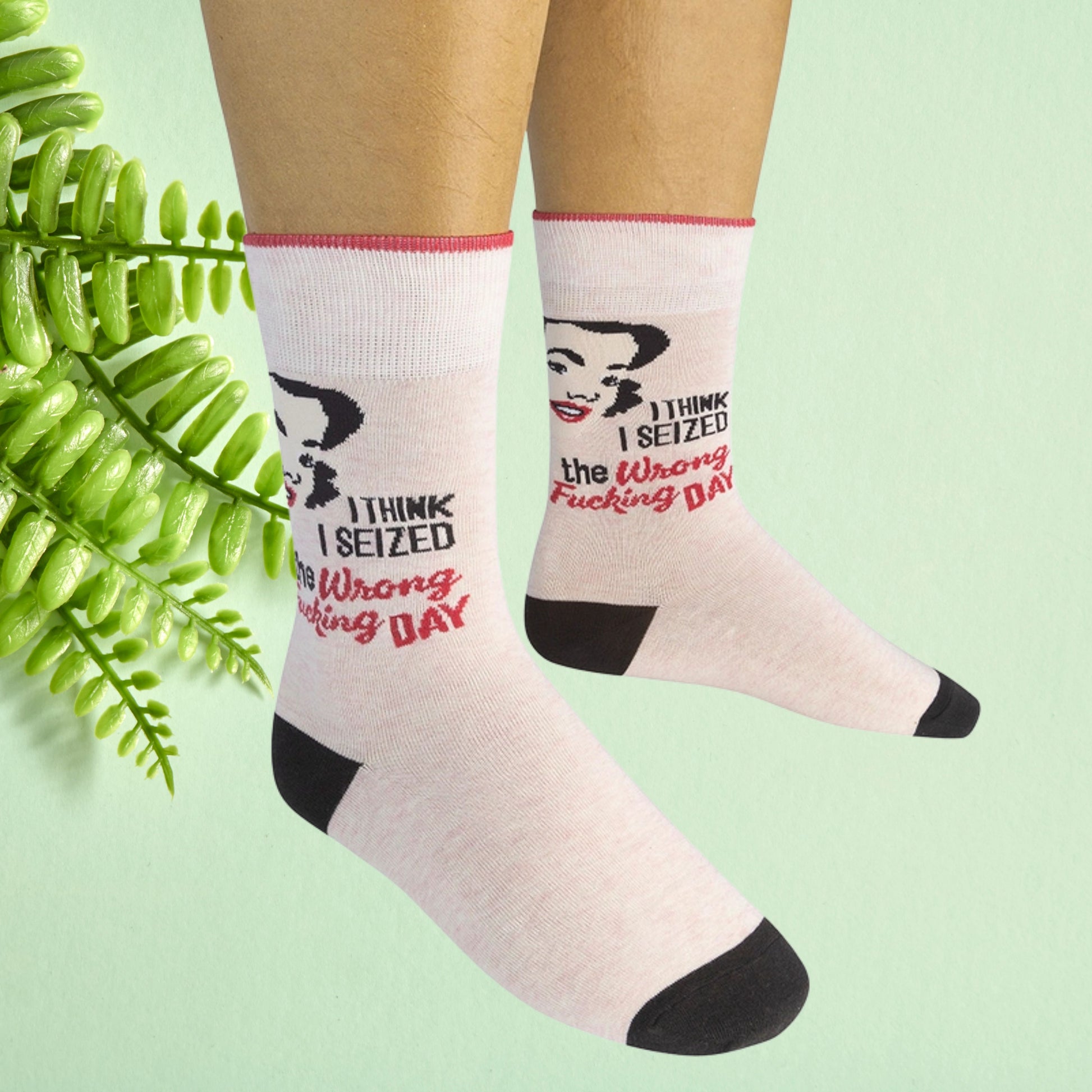 Cream crew socks with retro woman graphic and “I think I seized the wrong fucking day” text, worn on feet against a soft green background.