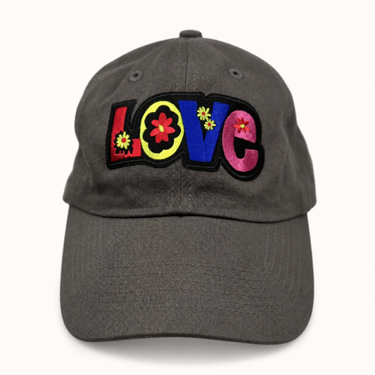 A dark gray baseball cap featuring a colorful embroidered 'LOVE' patch on the front, with each letter decorated with small flower details, set against a plain white background.