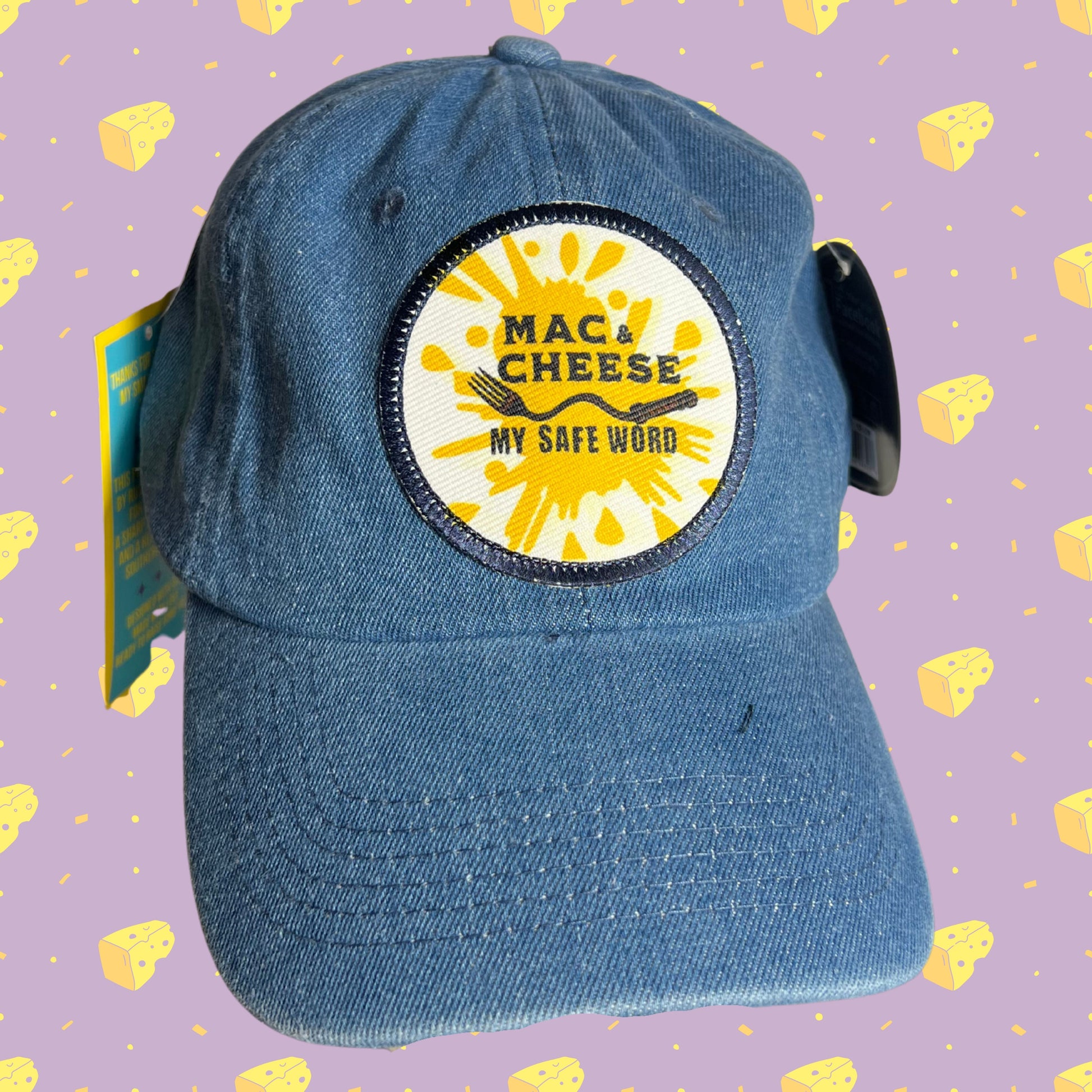 Denim embroidered patch hat with yellow splash design and bold text “Mac & Cheese My Safe Word,” displayed against purple background with repeating cheese icons.