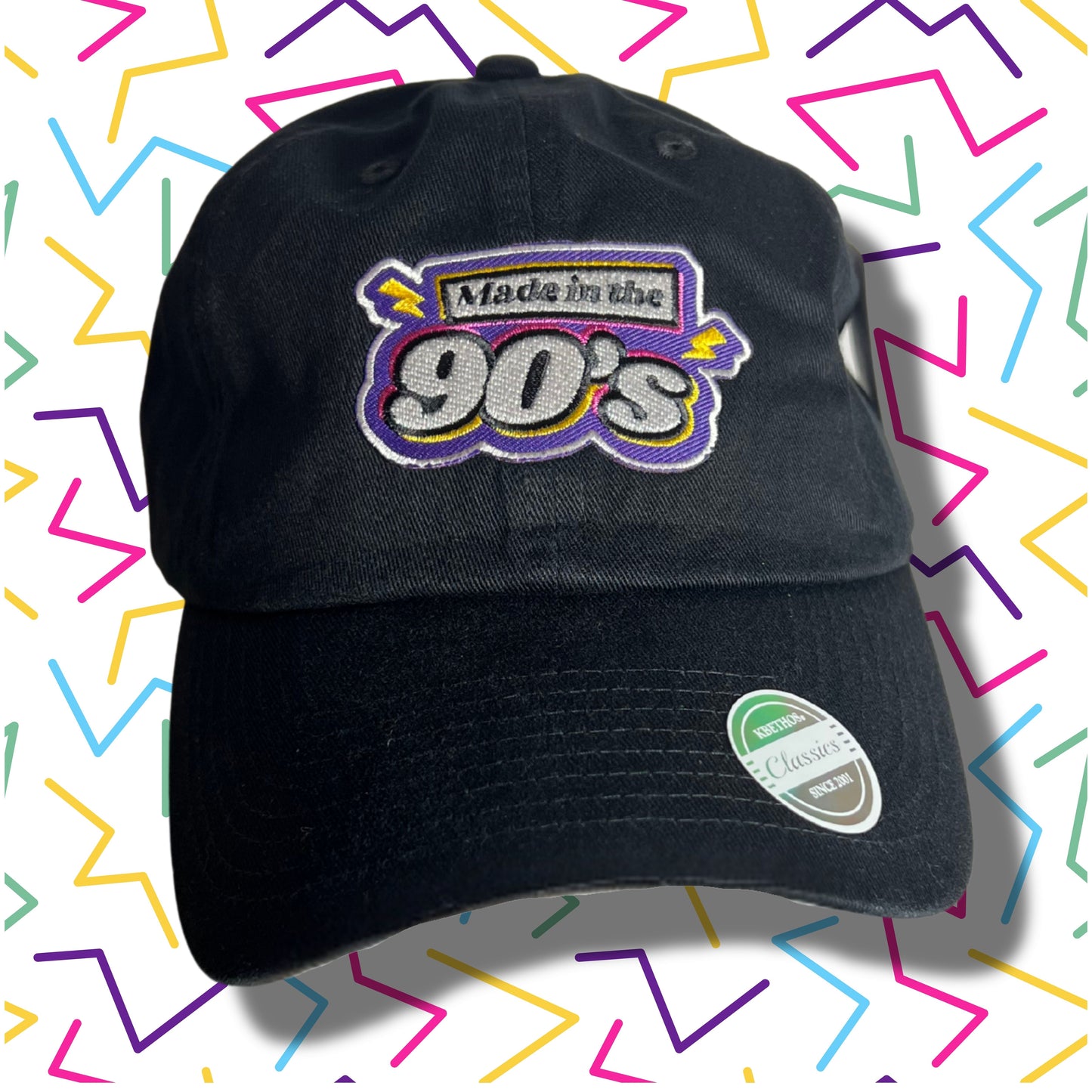 Black baseball cap with embroidered text “Made in the 90’s” in bold retro colors, displayed against a colorful zig-zag 90’s style background.