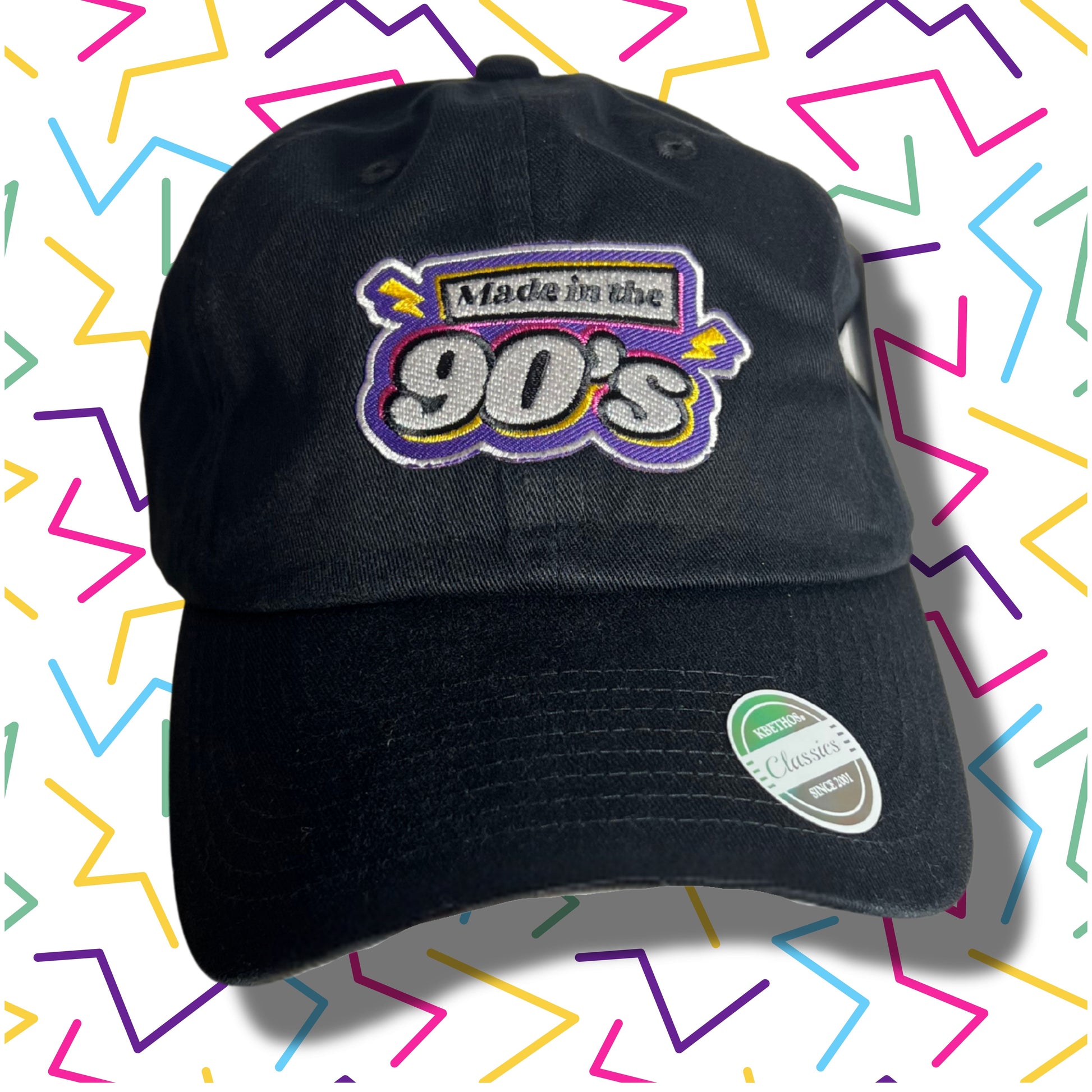 Black baseball cap with embroidered text “Made in the 90’s” in bold retro colors, displayed against a colorful zig-zag 90’s style background.