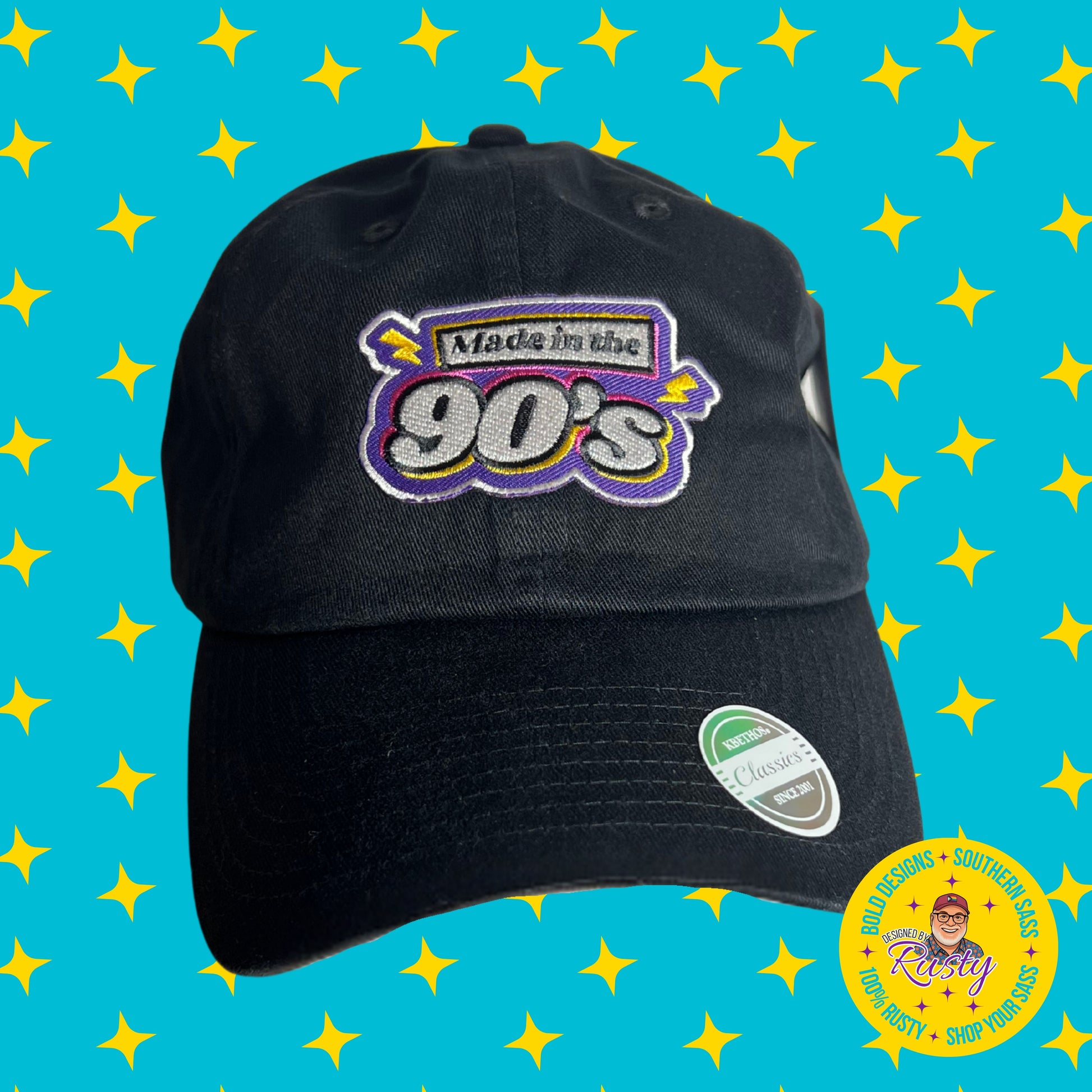 Black baseball cap with embroidered text “Made in the 90’s” in bold retro colors, on turquoise background with yellow stars and Designed by Rusty logo.