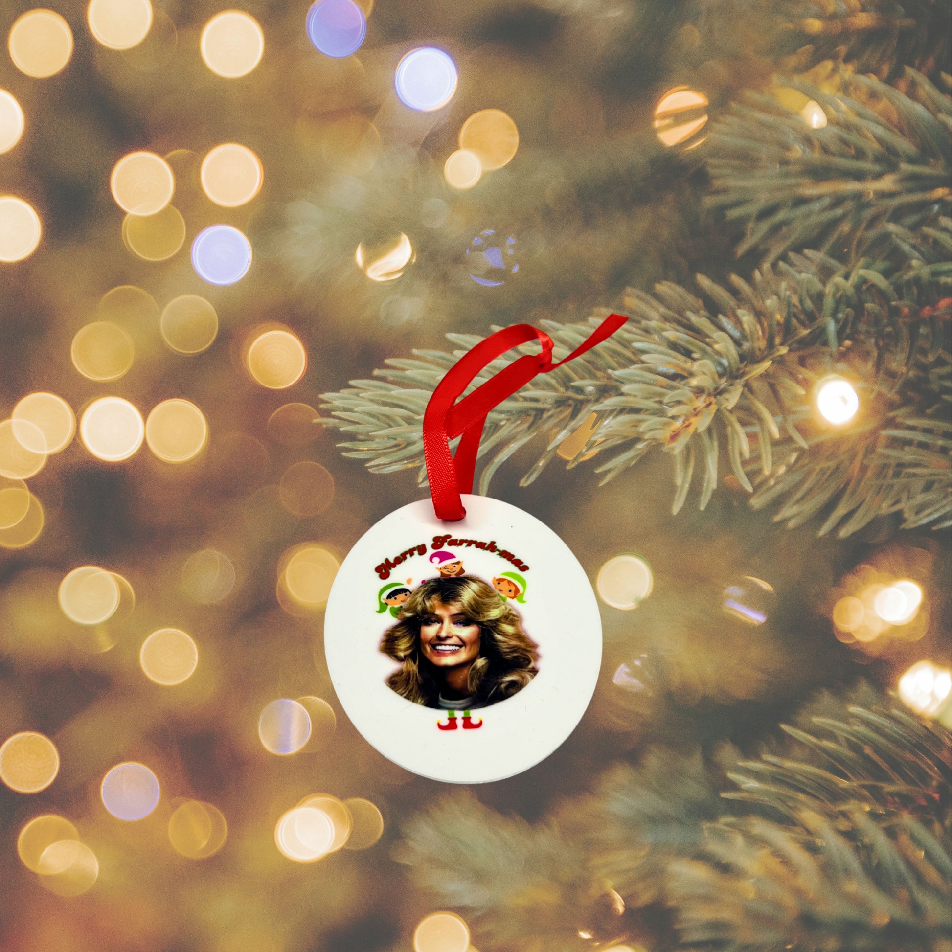 White ceramic “Merry Farrahmas” ornament featuring Farrah Fawcett and cartoon elves, hanging from a Christmas tree branch with warm holiday lights in the background.