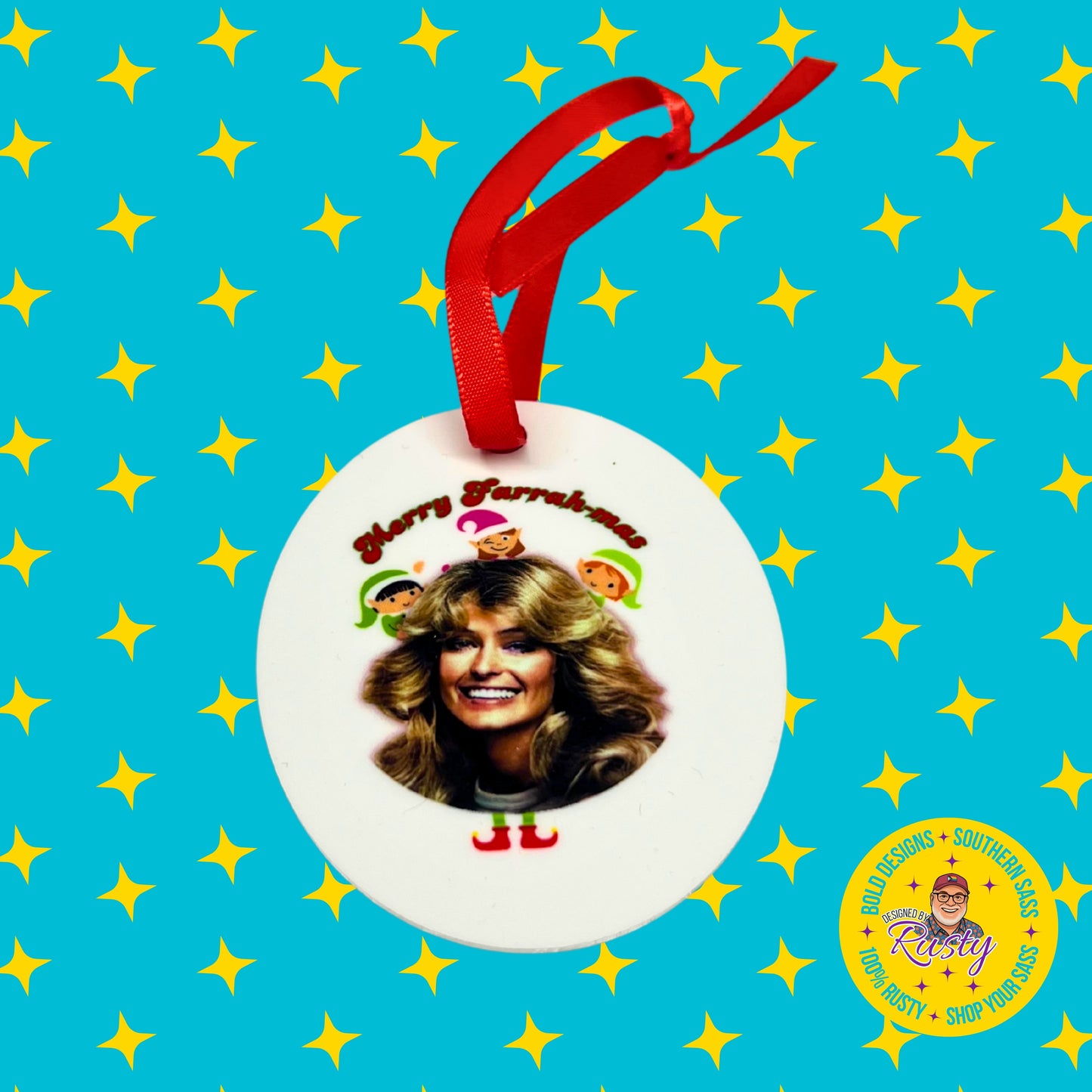 Designed by Rusty branded photo of a white ceramic “Merry Farrahmas” ornament featuring Farrah Fawcett and colorful elves, shown with a red hanging ribbon on a turquoise background with yellow stars.