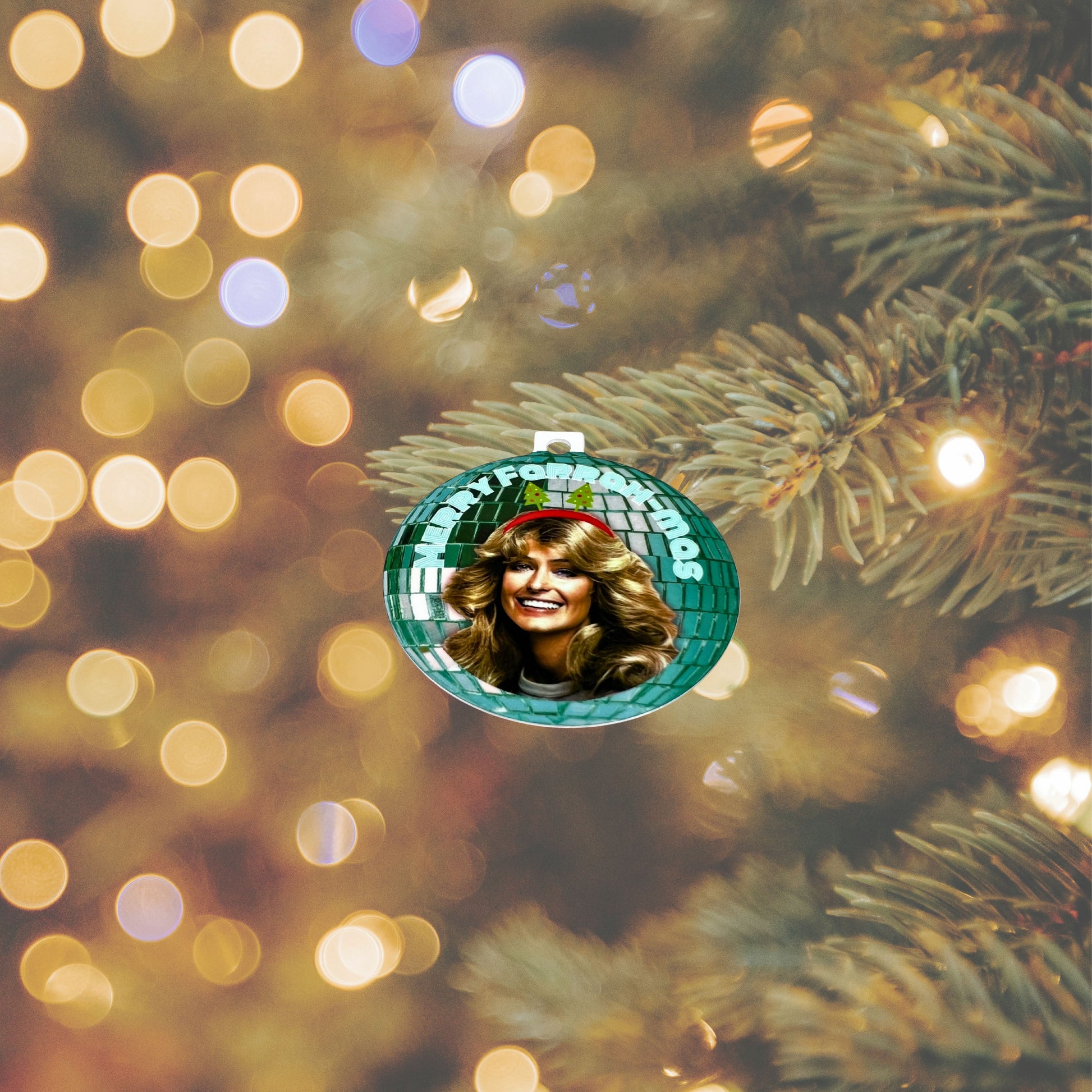 ‘Merry Farrah-mas’ Christmas ornament with Farrah Fawcett and green disco-ball design, hanging on a Christmas tree branch with warm holiday lights