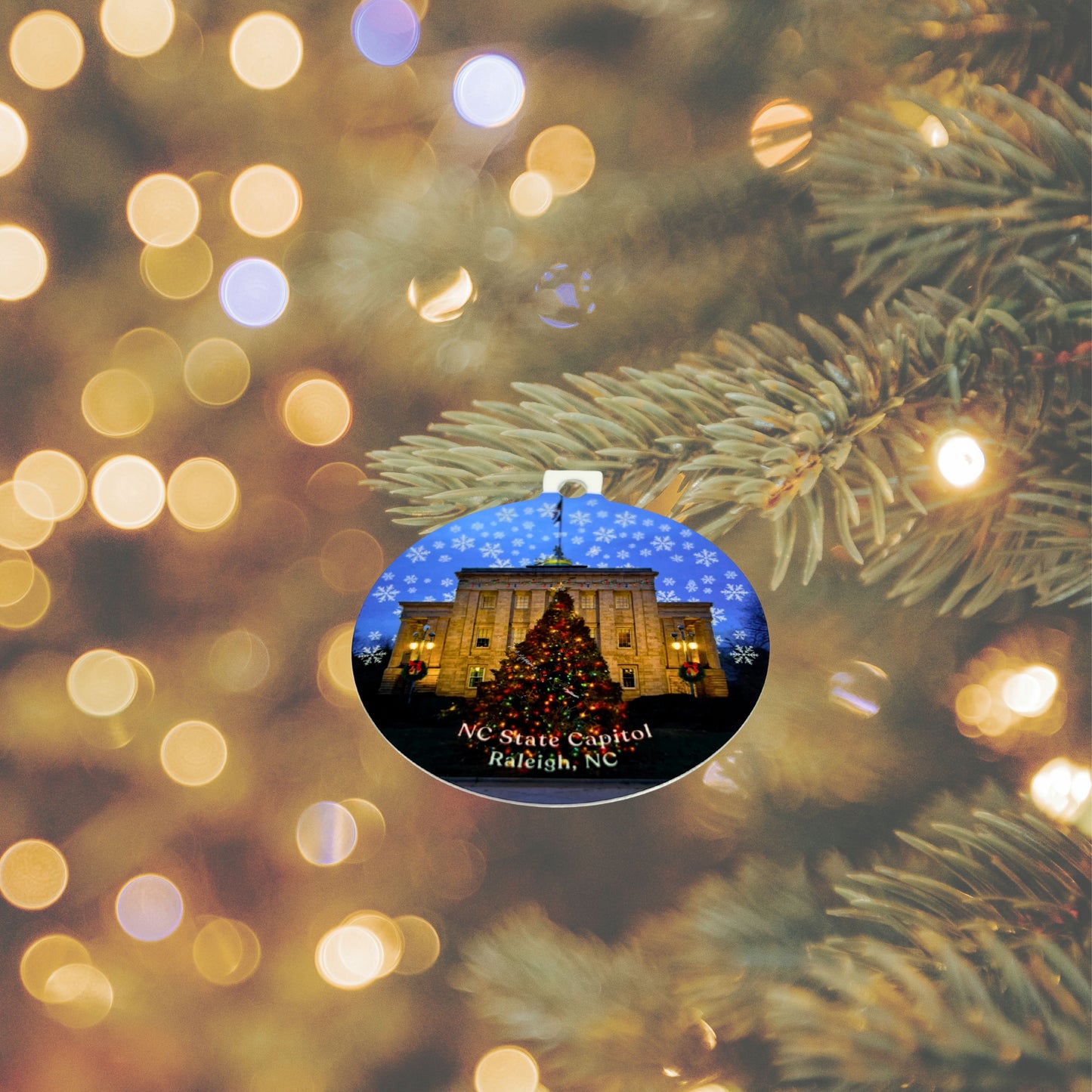 NC State Capitol Christmas ornament hanging on a lit Christmas tree, showing the State Capitol building and decorated tree with snowflakes.