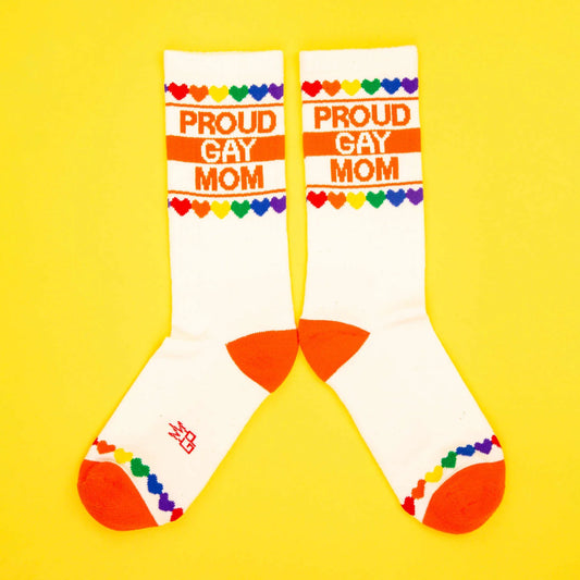 White novelty socks with orange accents, rainbow heart trim, and bold text: “Proud Gay Mom” on yellow background