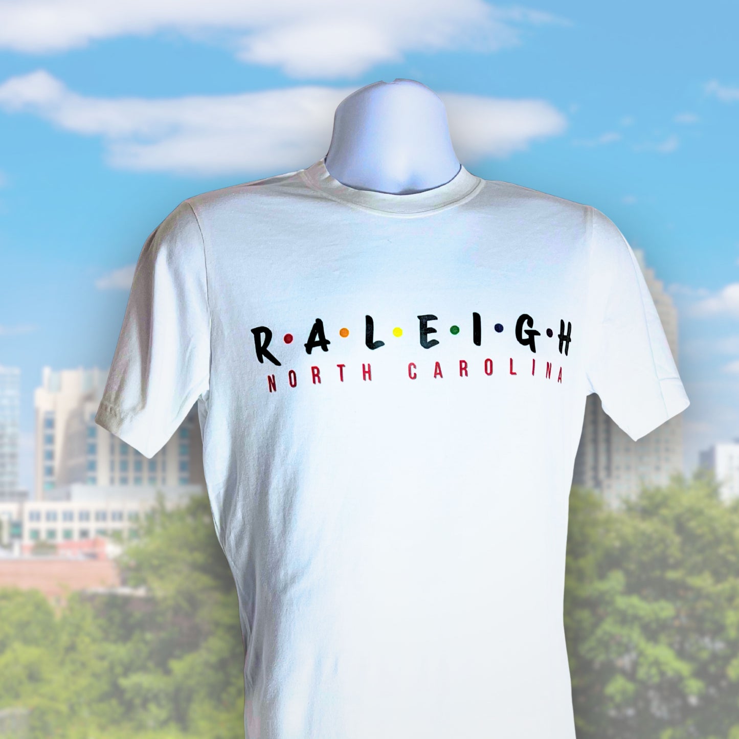 White t-shirt with 'RALEIGH NORTH CAROLINA' text on a mannequin against a blurred cityscape background