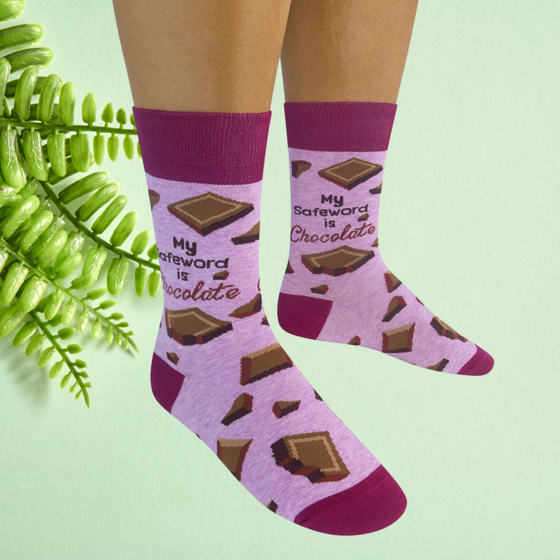Pink crew socks with chocolate bar pattern and “My safeword is chocolate” text worn on feet against a soft green background.