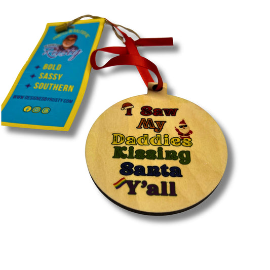 Round wooden Christmas ornament with colorful text reading ‘I Saw My Daddies Kissing Santa Y’all,’ tied with a red ribbon.