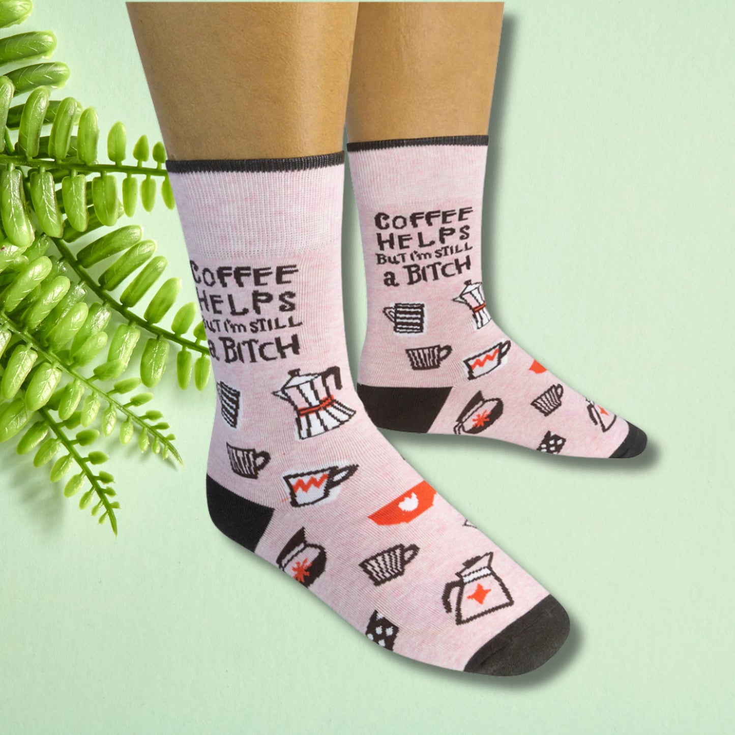Light pink crew socks with coffee illustrations and “Coffee helps but I’m still a bitch” text, worn on feet against a soft green background.