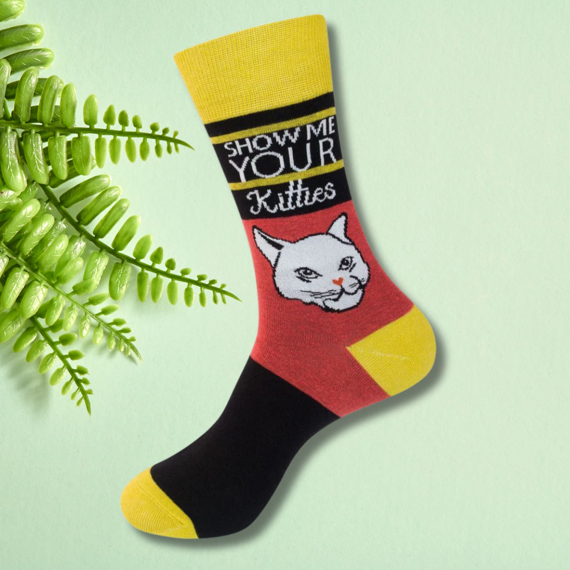 Colorblock crew socks with cat illustration and “Show me your kitties” text, worn on feet against a soft green background.