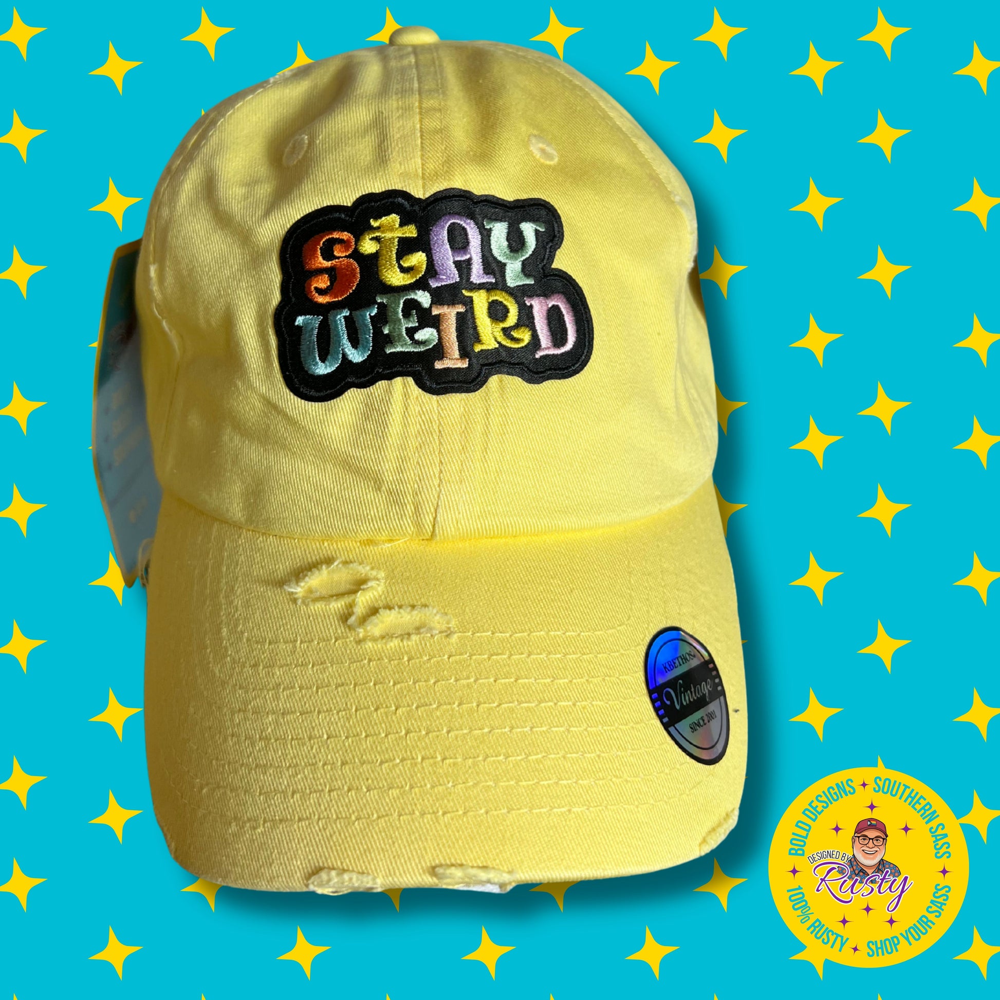 Yellow embroidered patch hat with multicolor “Stay Weird” text, on turquoise background with yellow stars and Designed by Rusty logo.