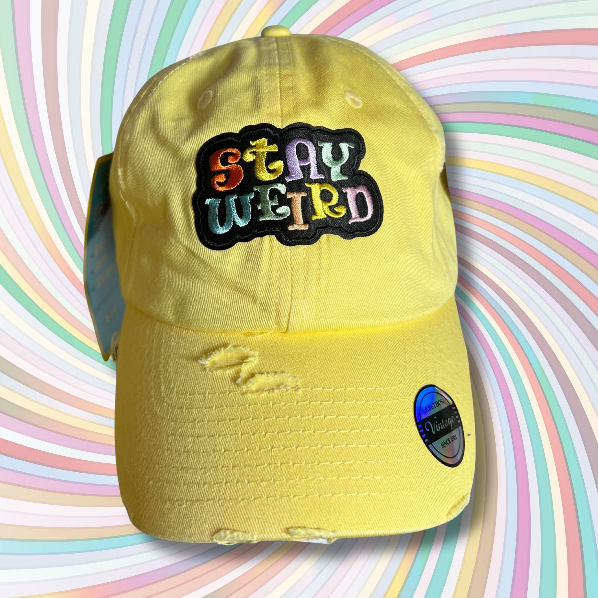 Yellow embroidered patch hat with multicolor “Stay Weird” text, displayed against retro swirl rainbow background.
