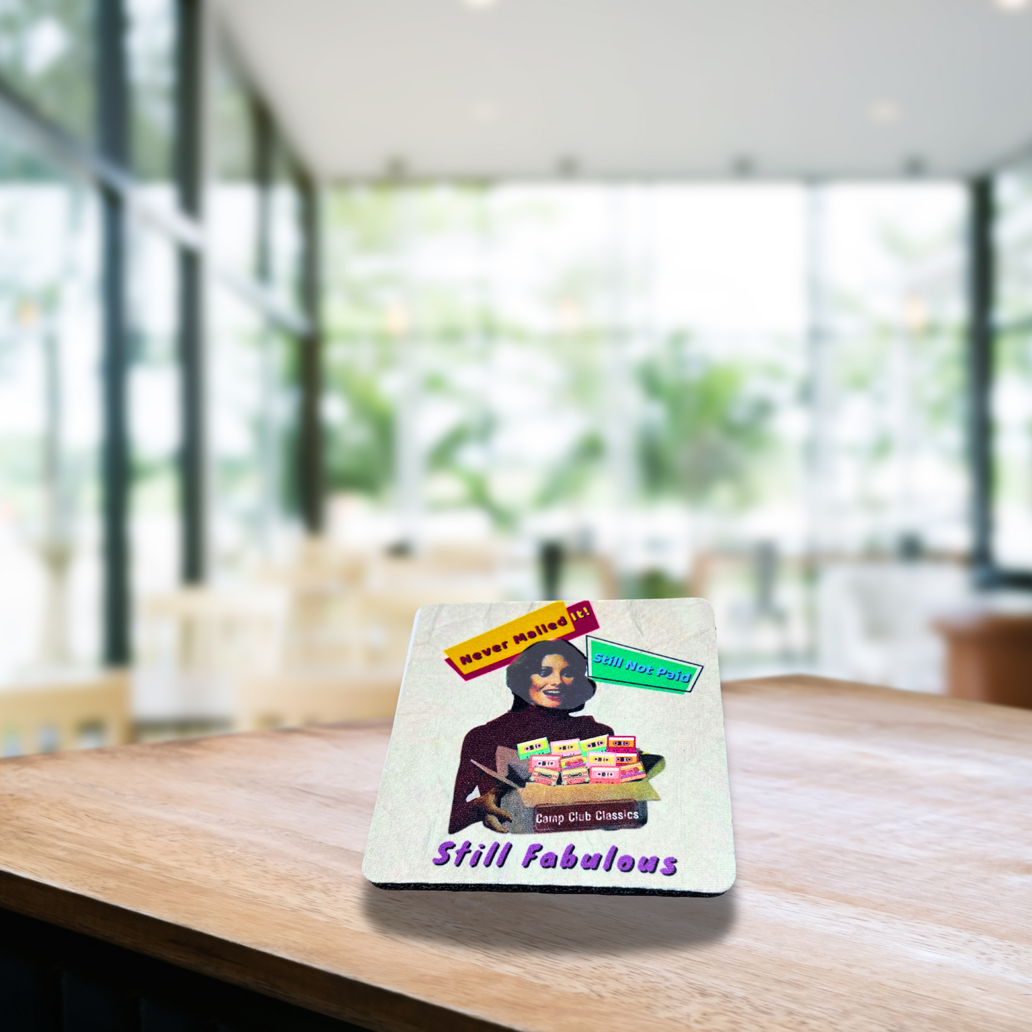 White 4x4 coaster with rainbow “Still Fabulous” design, displayed on a natural wood table.