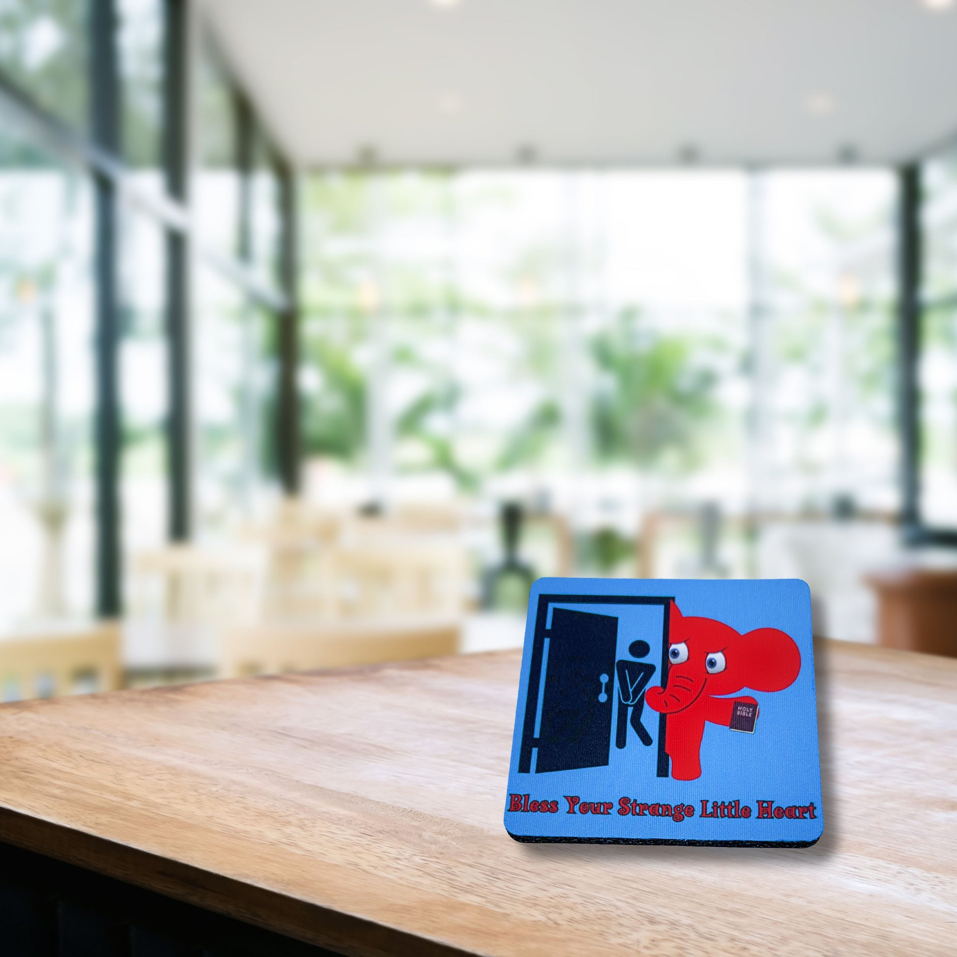 Humorous coaster reading “Bless Your Strange Little Heart” with red elephant design, displayed on wooden café table in bright modern setting.