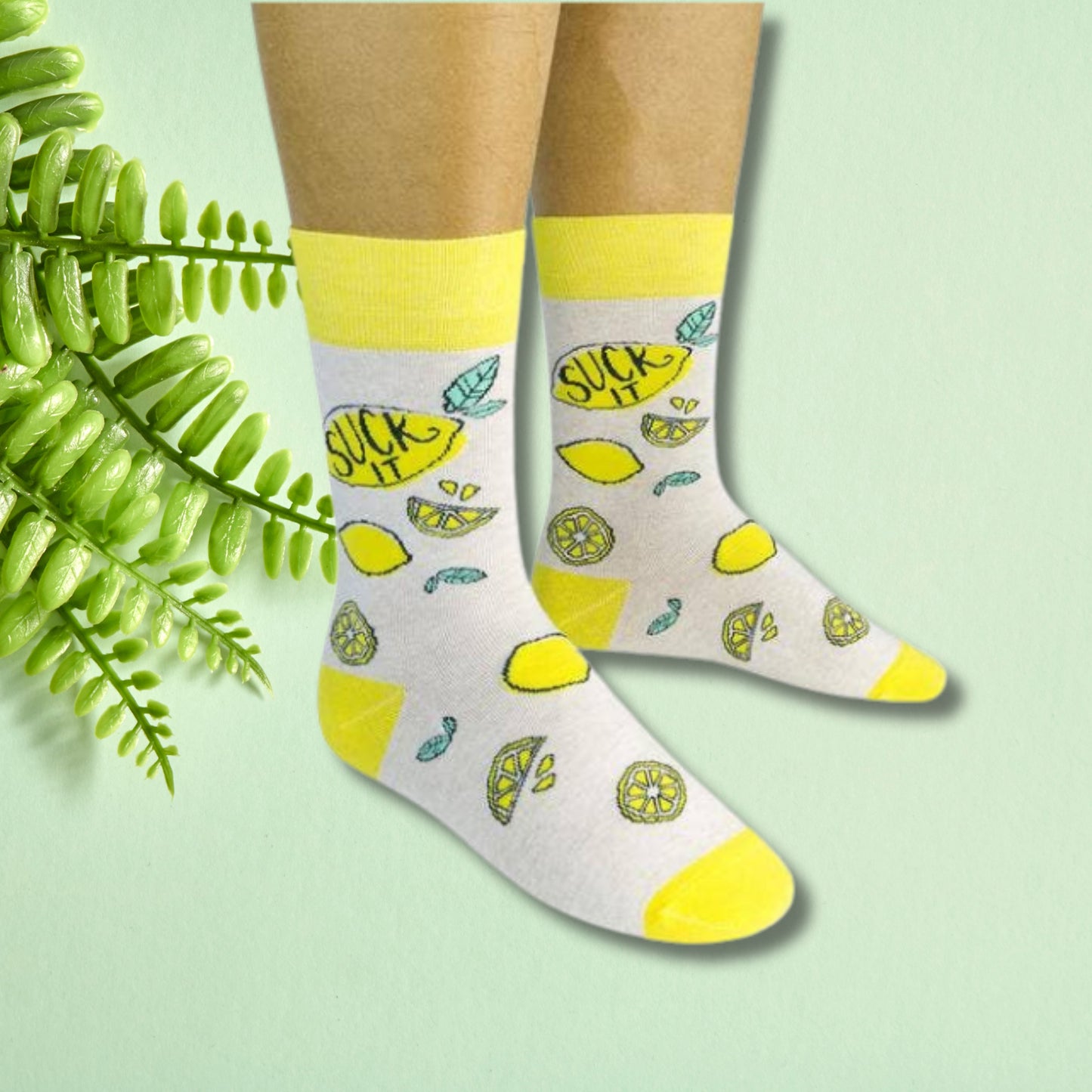 Gray and yellow crew socks with lemon illustrations and “Suck it” text worn on feet against a soft green background.