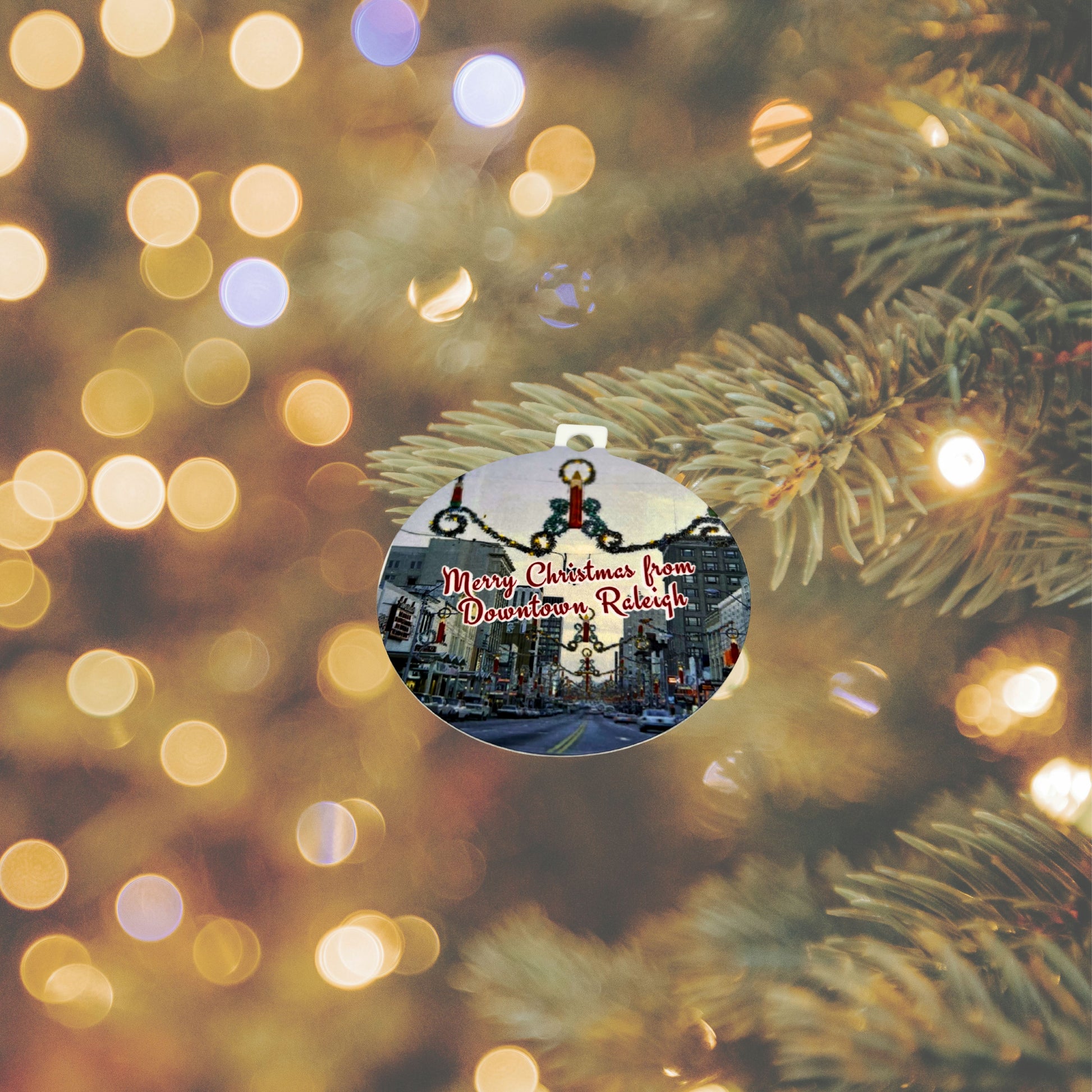 Round aluminum ornament printed with a vintage Downtown Raleigh Christmas scene hanging on a Christmas tree branch with warm golden lights in the background.”