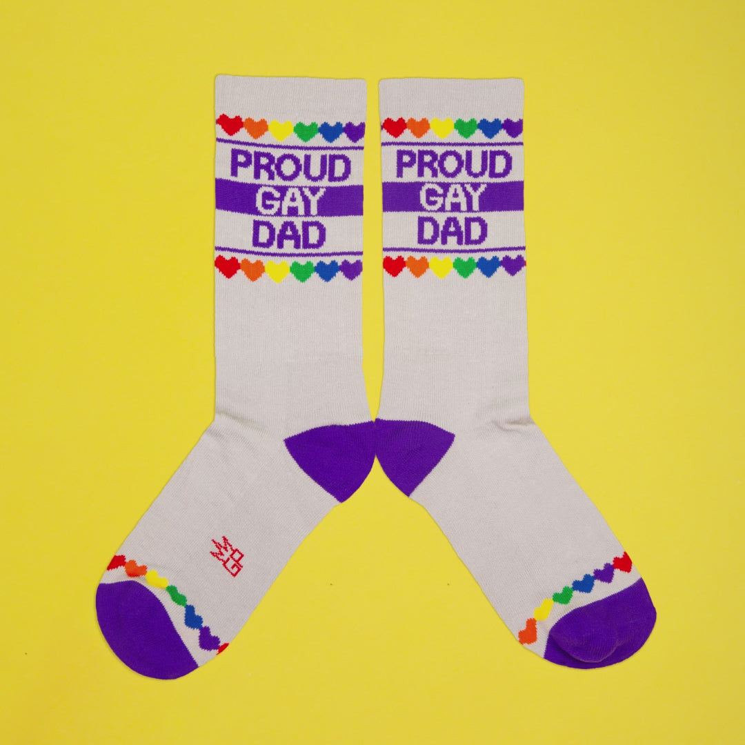grayish crew socks with purple heel and toe, rainbow heart trim, and bold text: “Proud Gay Dad” on yellow background